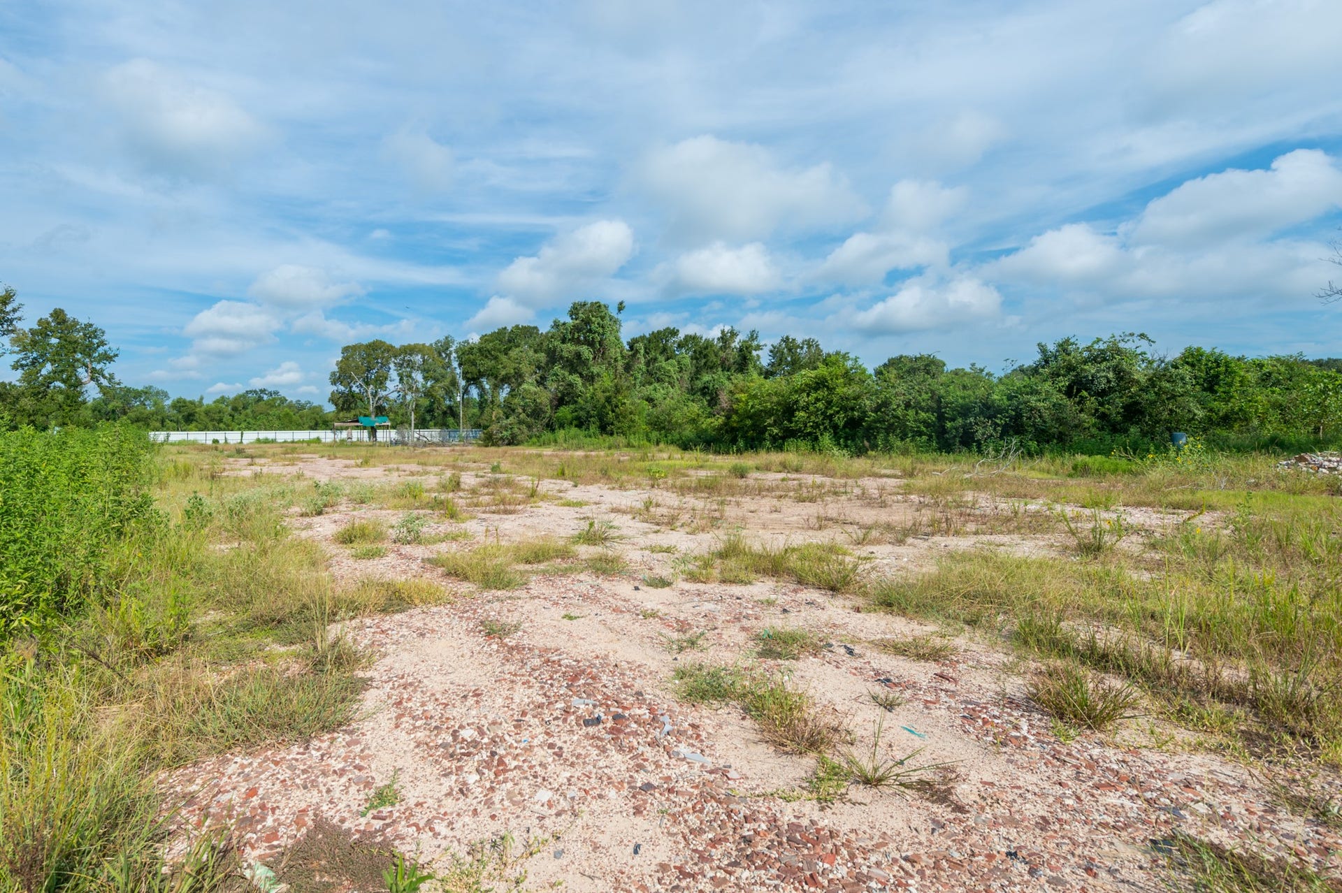 Expansive vacant lot with a mix of grass and sand, surrounded by lush greenery. Ideal for development, offering a serene natural setting under a clear blue sky.