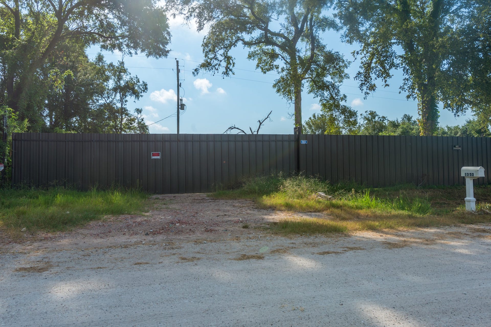 This photo features a gated property entrance with a large metal fence, offering privacy. The surrounding area is lush with trees, and there's a dirt driveway leading to the gate. A mailbox is visible, indicating a residential setting.