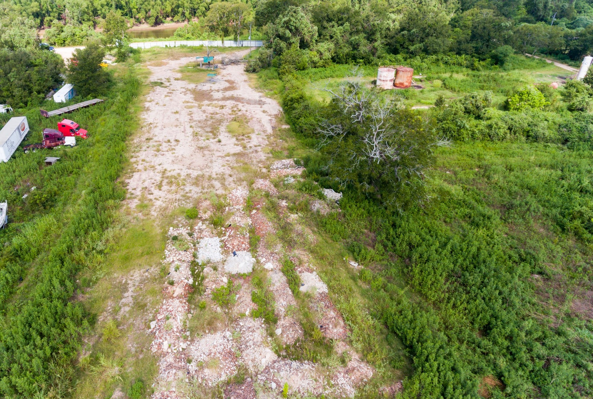 This photo shows a large, open plot of land with some overgrown vegetation and scattered debris. There are a few trucks and trailers parked on one side, and a mix of grass and dirt covers the area. It is bordered by trees, offering a semi-rural setting, and could be a potential site for development or storage.