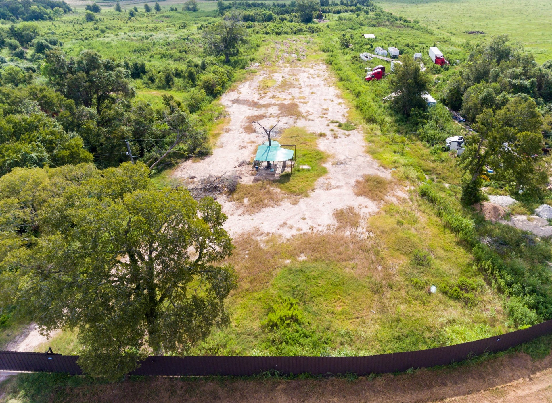 This photo shows a large, open plot of land with partial clearing, surrounded by lush greenery. There is a small structure in the center and a few vehicles and trailers in the distance, ideal for development or agricultural use.