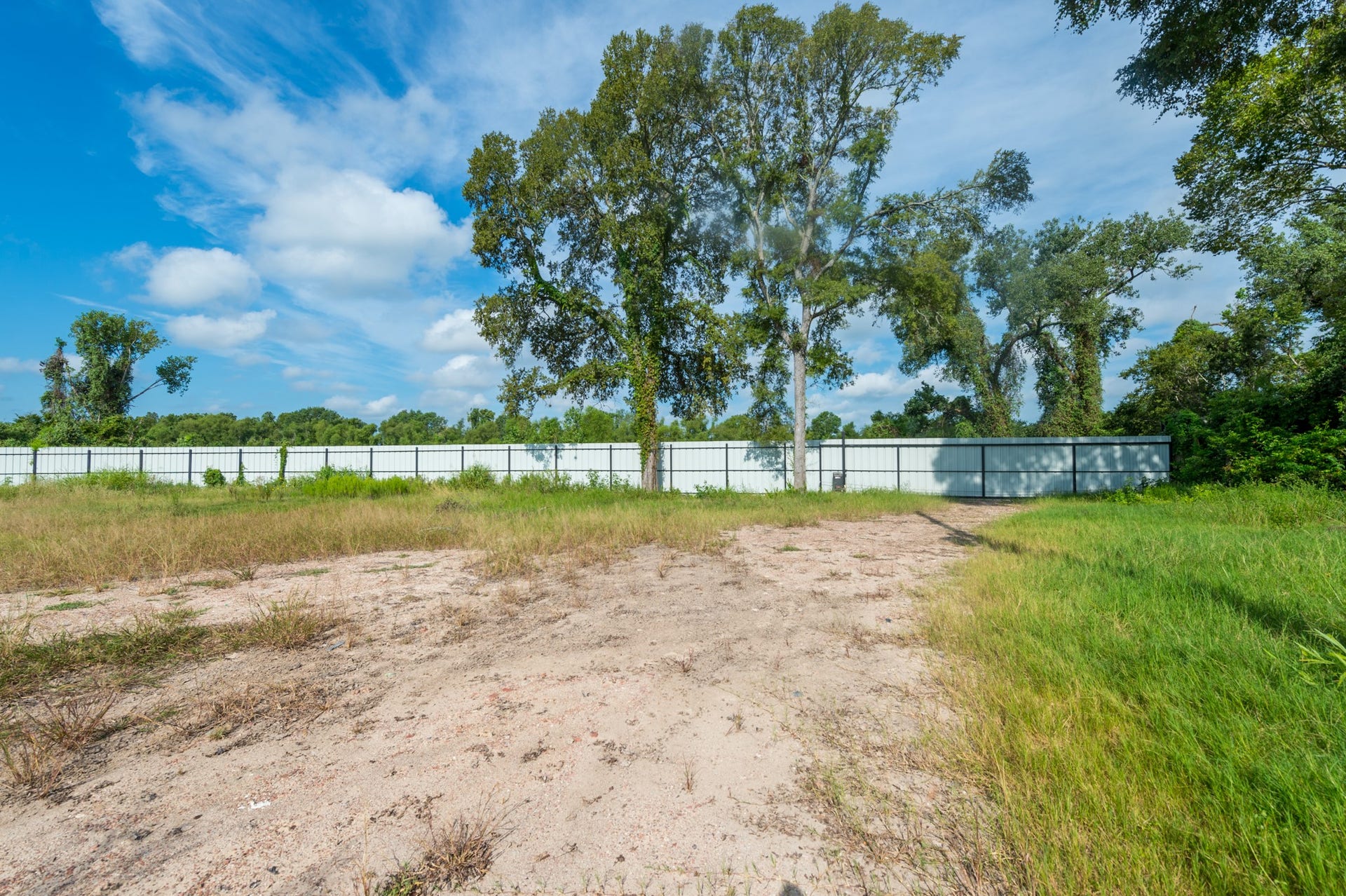 This photo shows a spacious, flat plot of land with a mix of grass and sandy soil, bordered by a tall white fence and surrounded by lush trees, perfect for building a private home.