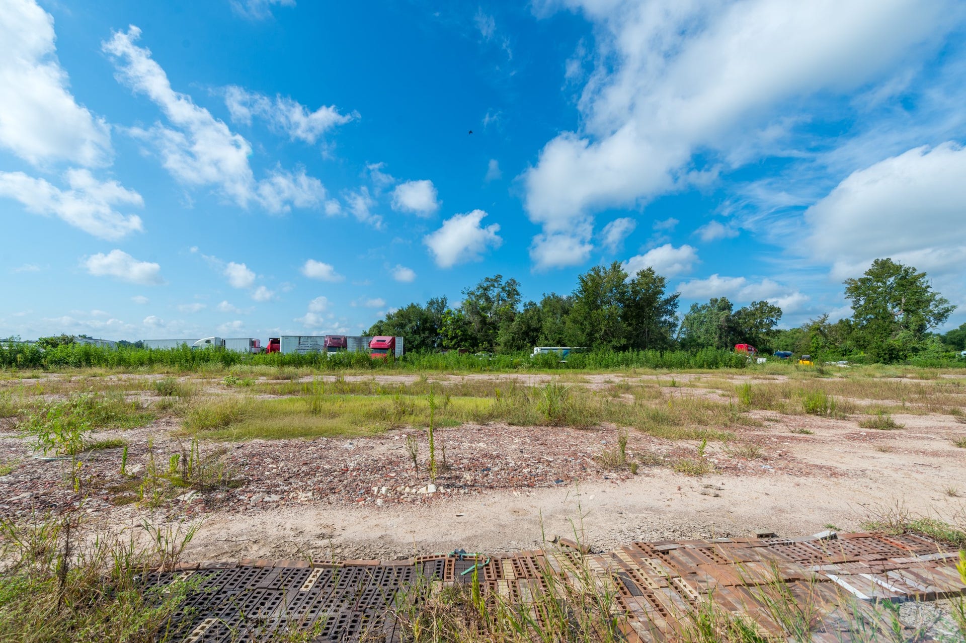 This photo shows an open plot of land with some sparse grass and trees in the background. The sky is clear with a few clouds, suggesting a sunny day. It's a spacious area, possibly suitable for development or landscaping.