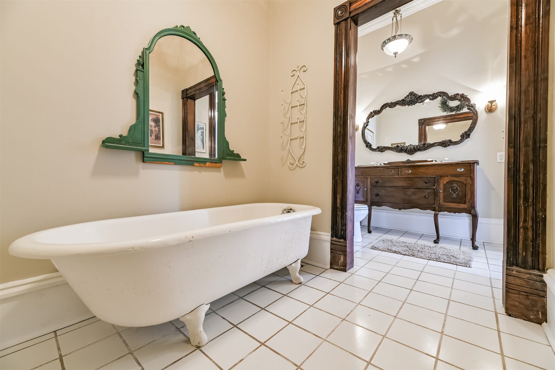 1927 Broadway Street, Galveston, TX 77550 This charming bathroom services the secondary bedrooms, featuring a dresser-style vanity with dual basin sinks, a period-specific ornately framed mirror, tile floors, and a separate room with a cast iron clawfoot bathtub.