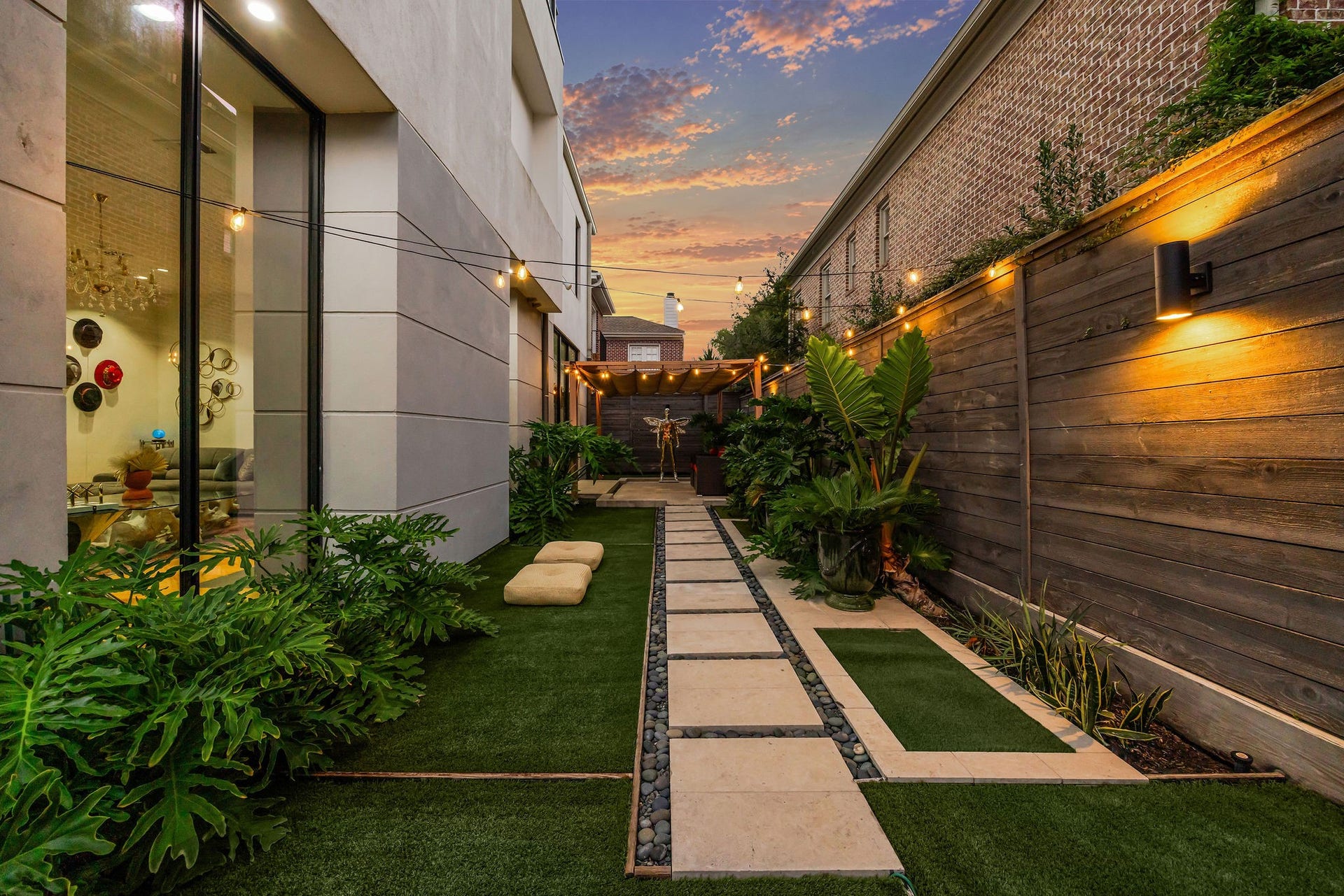 1310 Nantucket Drive, Houston, TX 77057 Experience serene outdoor living in this beautifully landscaped backyard, featuring a modern walkway, lush greenery, and ambient lighting for a tranquil atmosphere.