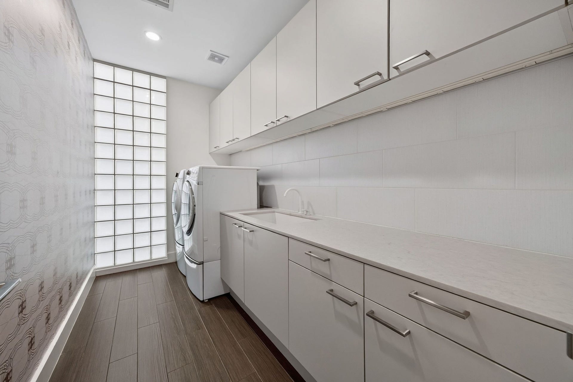 1310 Nantucket Drive, Houston, TX 77057 Sleek and modern laundry room featuring abundant cabinetry, built-in washer and dryer, a convenient sink, and stylish glass block wall for natural light.