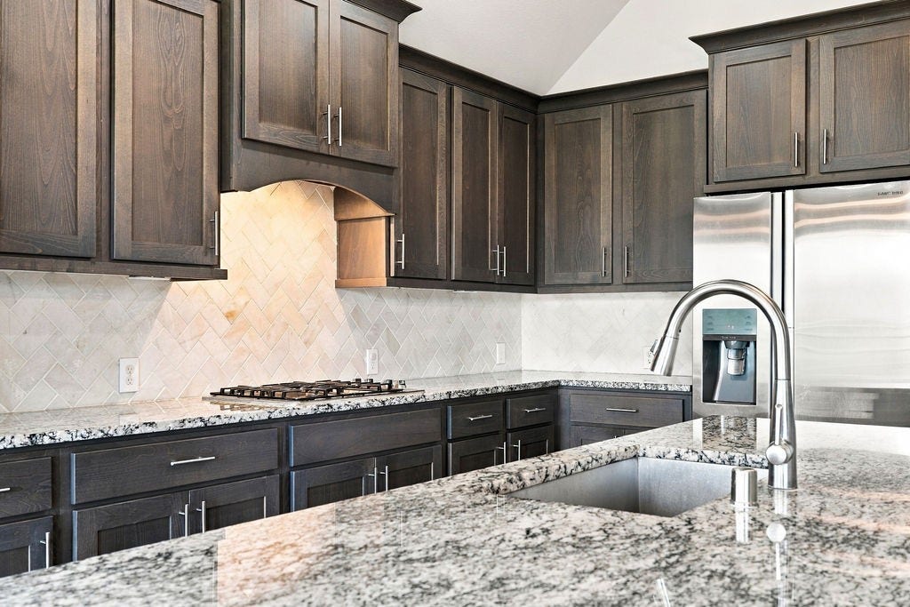 6914 Red Oak Drive, Katy, TX 77493 Elegant kitchen with dark wood cabinetry, granite countertops, and a sleek stainless steel refrigerator, complemented by a modern faucet and a stylish tile backsplash.