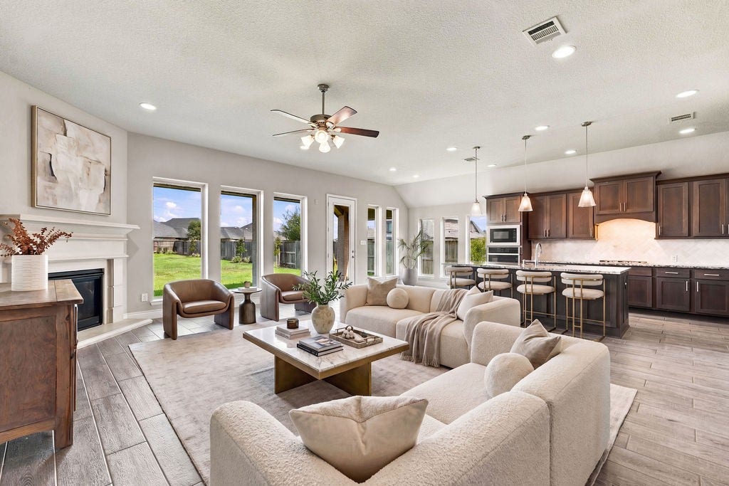 6914 Red Oak Drive, Katy, TX 77493 Spacious open-concept living area featuring a cozy fireplace, elegant wood flooring, and a modern kitchen with dark cabinetry and a large island. Natural light floods through multiple windows, enhancing the warm, inviting atmosphere.