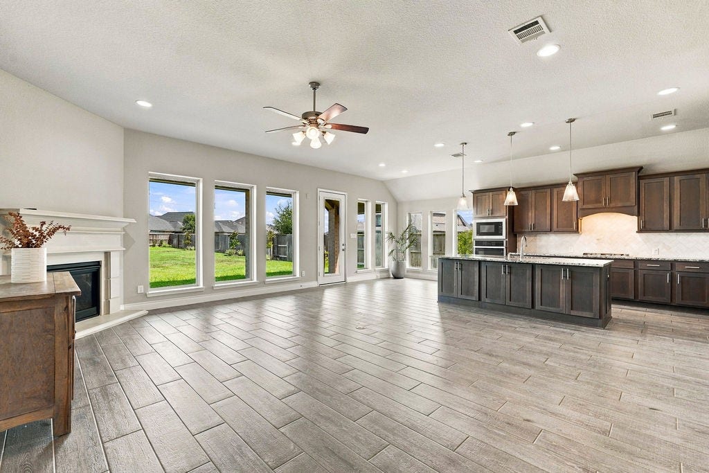 6914 Red Oak Drive, Katy, TX 77493 Spacious open-concept living area with elegant wood-look tile flooring, large windows offering abundant natural light, and a modern kitchen featuring dark cabinetry and a central island.