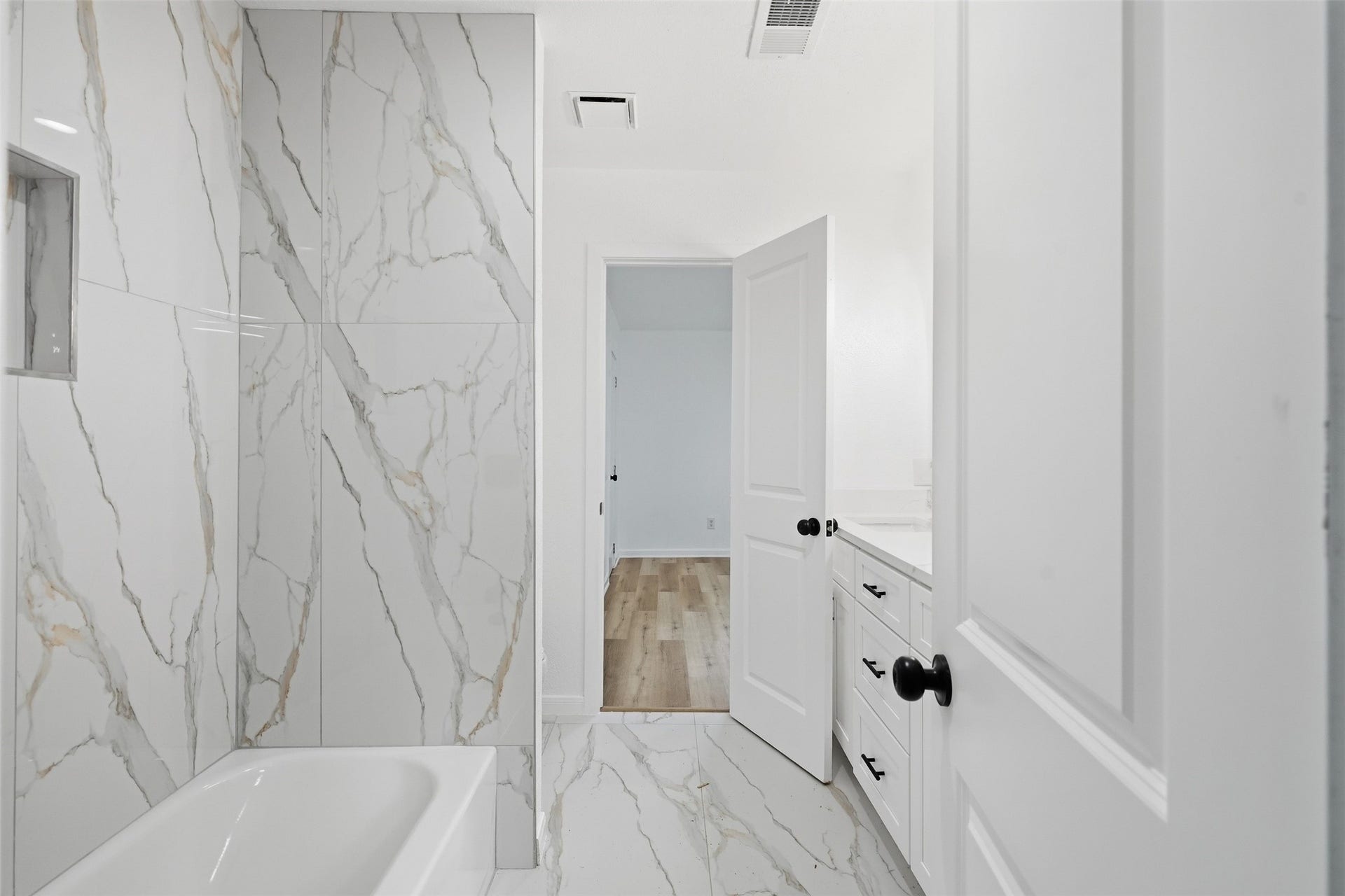 1936 Upland Drive, Houston, TX 77043 Sleek bathroom featuring elegant marble-patterned tiles, a modern bathtub, and stylish cabinetry, leading into a bright, airy room with wood flooring.