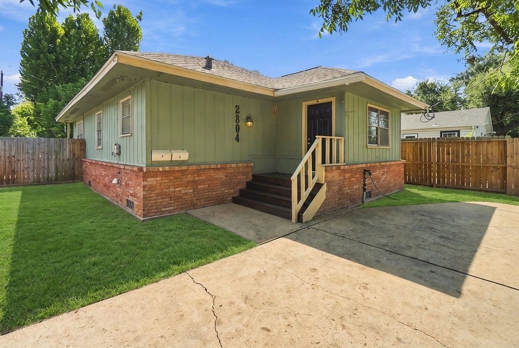 Charming duplex with a cozy porch, brick accents, and a spacious driveway, surrounded by a well-maintained fenced yard.