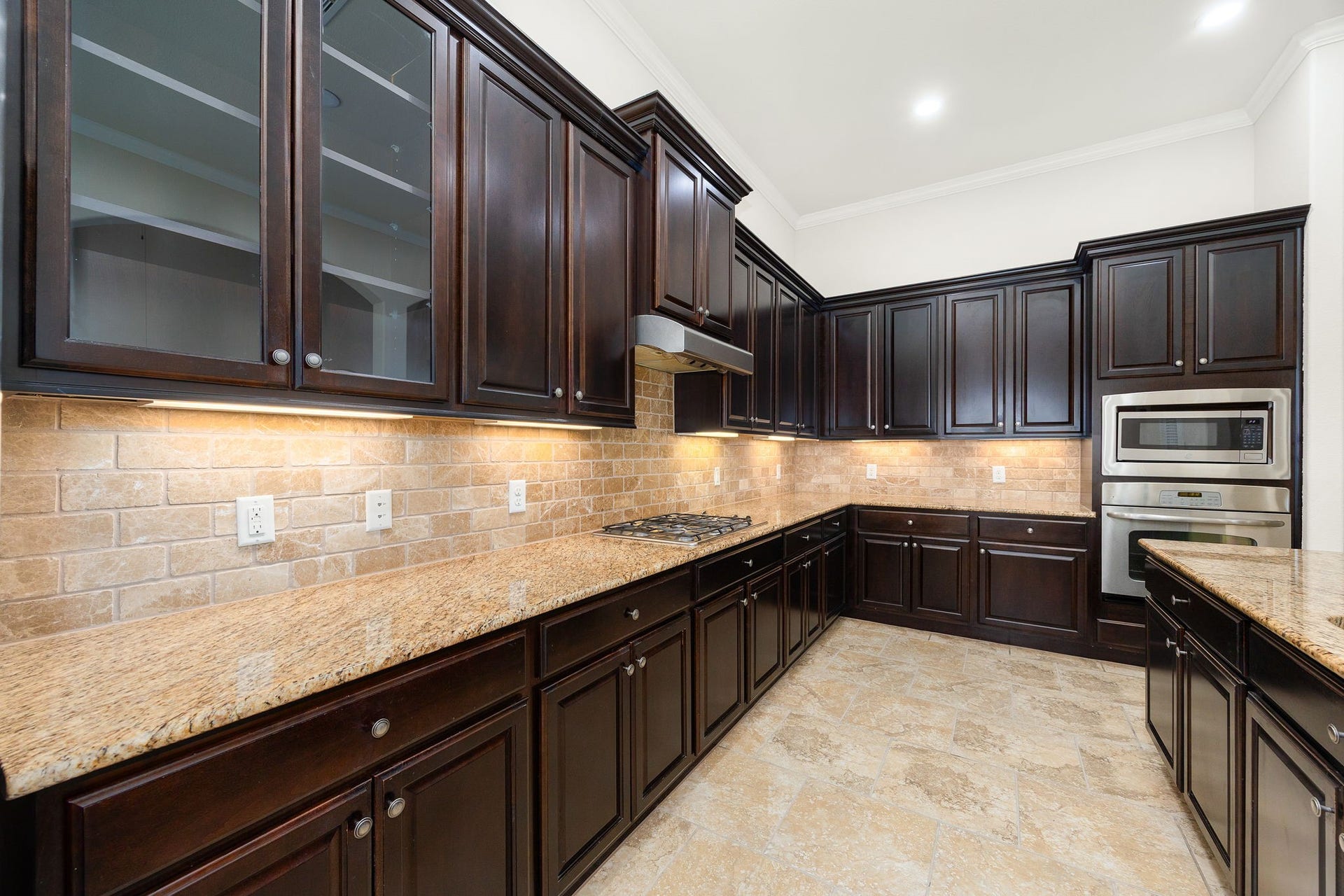 9931 Hutton Park Drive, Katy, TX 77494 The heart of the home showcases elegant glass-front display cabinetry, extensive under-cabinet accent lighting, and premium built-in appliances. This closer angle highlights the seamless flow of the rich dark cabinetry paired beautifully with the polished granite countertops. Complete with a built-in microwave, wall oven, and sleek cooktop, this thoughtfully designed layout maximizes both storage and prep space.