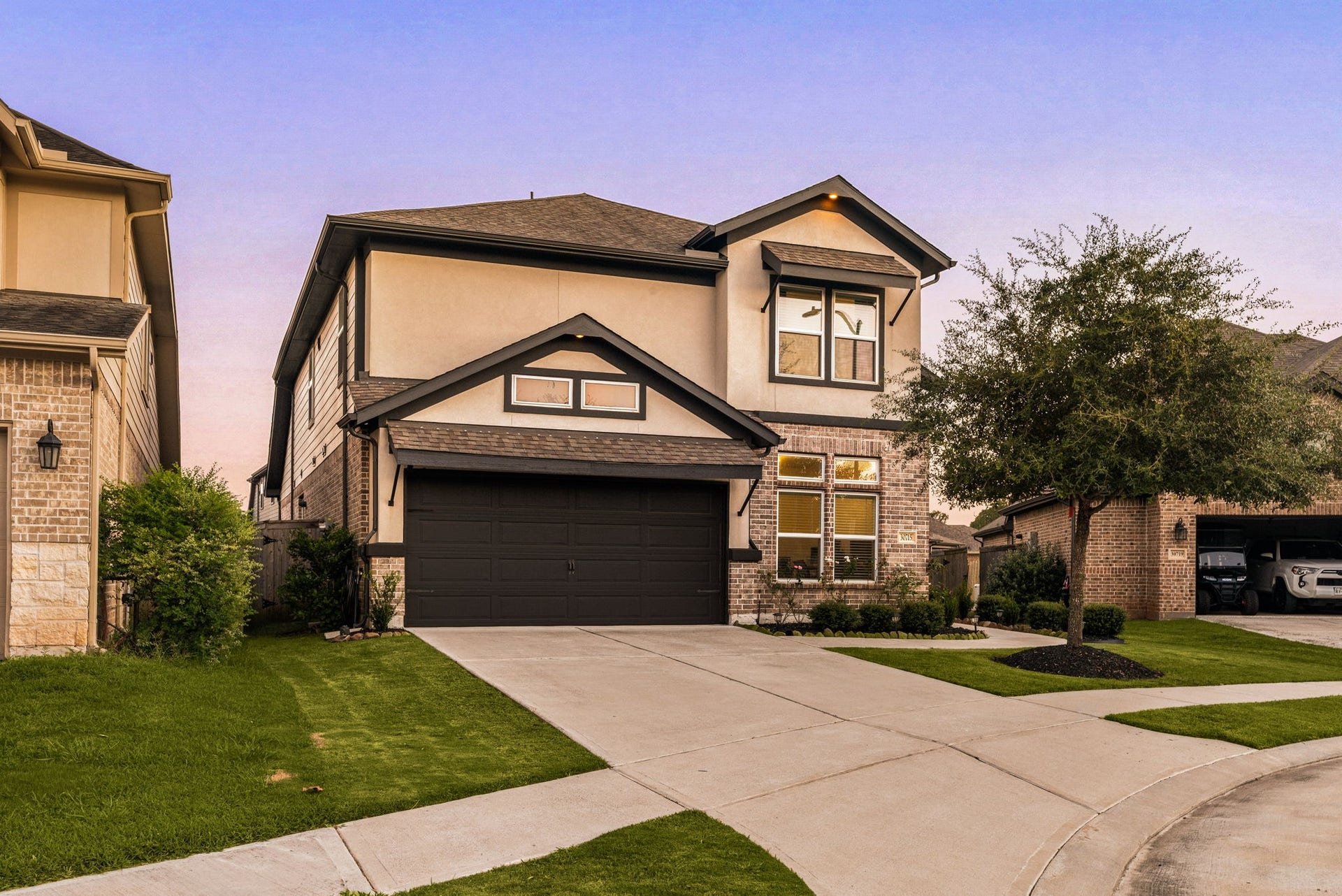 30715 Parkside Passage Drive, Fulshear, TX 77441 The front elevation showcases an elegant blend of brick, smooth stucco, and striking dark trim accents. A wide, pristine driveway leads to the front-entry garage, framed by manicured lawns and a beautifully established shade tree.
