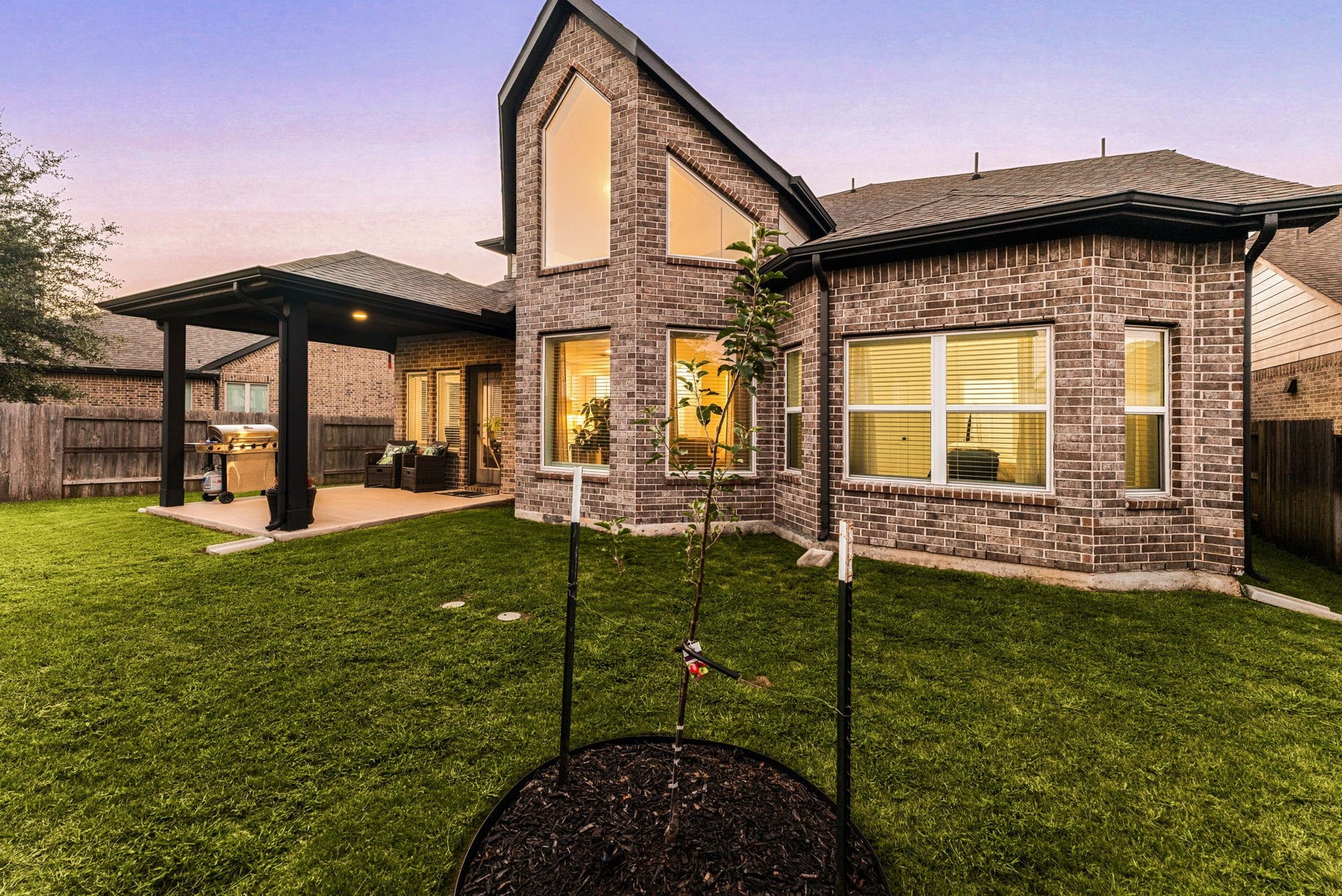 30715 Parkside Passage Drive, Fulshear, TX 77441 An outdoor oasis awaits in the sprawling, fully fenced backyard, anchored by an impressive covered patio pavilion with dark-toned support pillars perfect for open-air dining and lounging. The rear elevation showcases the dramatic, architectural gabled peak with soaring multi-pane clerestory windows.