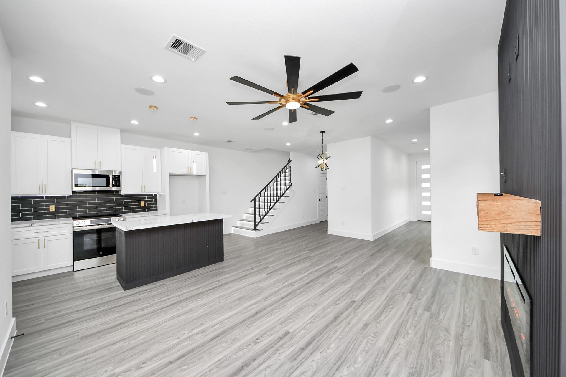 6711 Crestmont Street, Houston, TX 77033 Modern open-concept living area featuring sleek gray flooring, a stylish kitchen with a large island, and contemporary black backsplash. A striking ceiling fan and recessed lighting enhance the space, complemented by a chic staircase leading to the upper level.