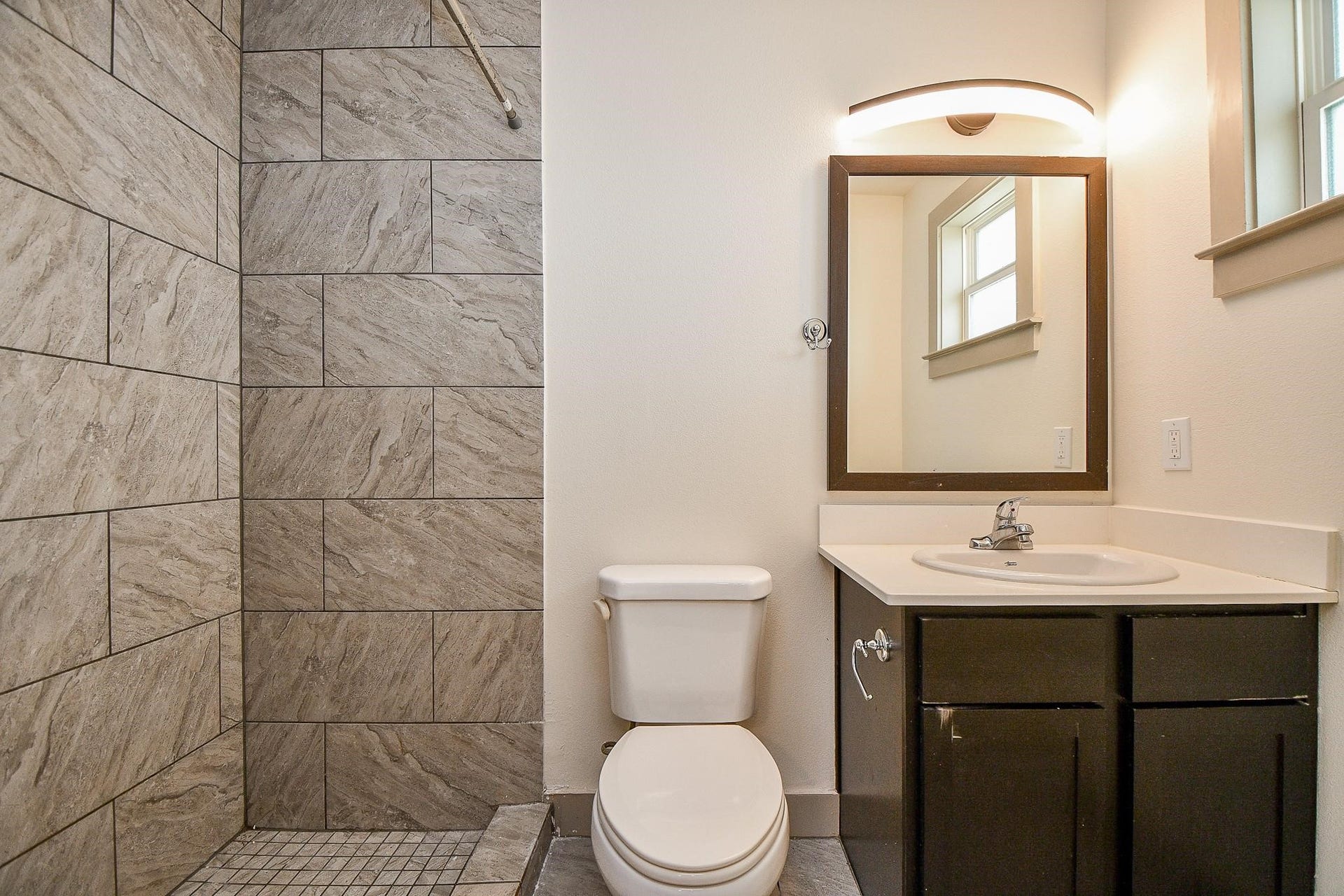 Modern bathroom with large gray tiled shower walls, white toilet, and sleek dark wood vanity with sink and overhead lighting.