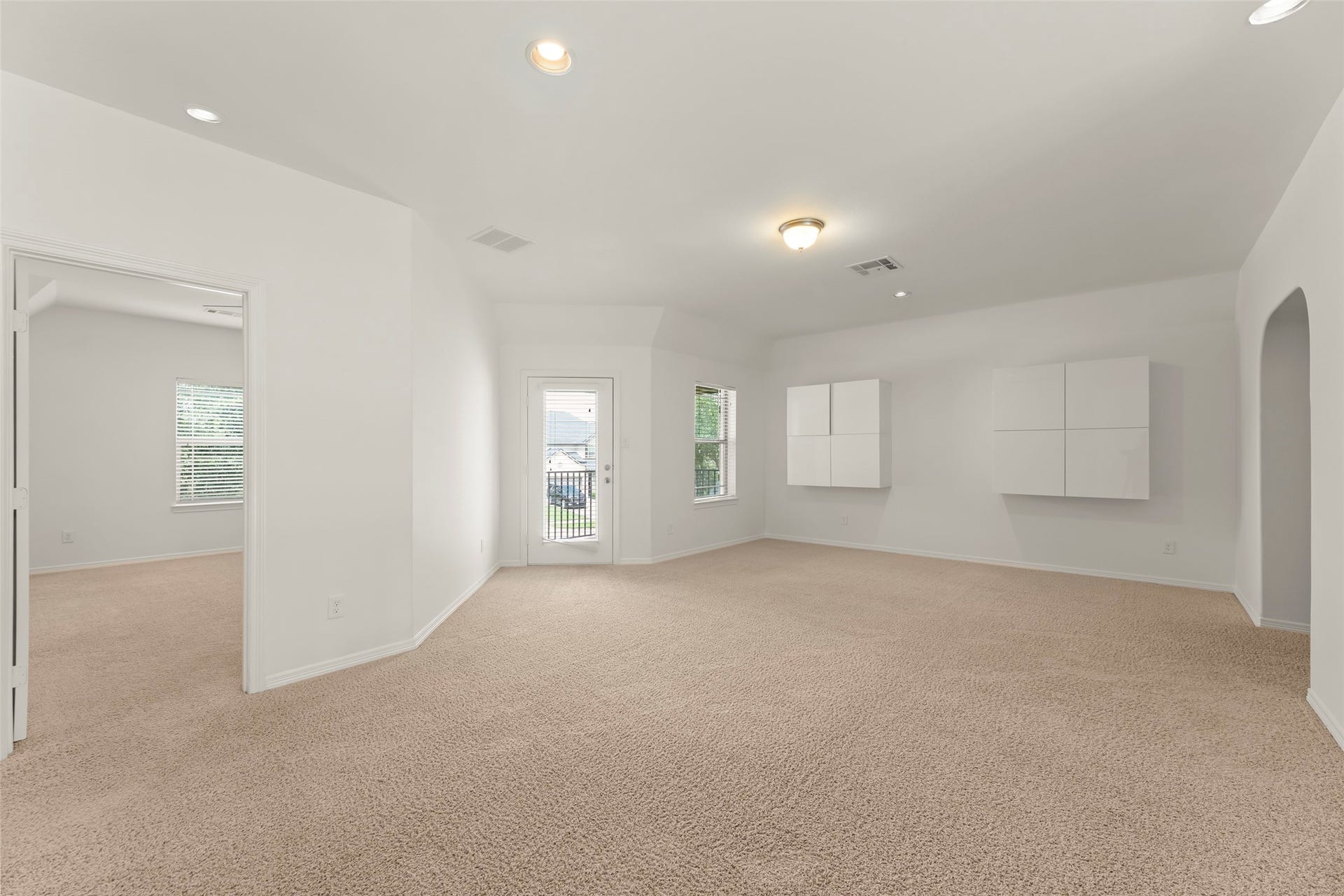 Spacious and bright room with plush carpeting, modern wall cabinets, and large windows offering ample natural light, leading to a serene outdoor view through a glass door.