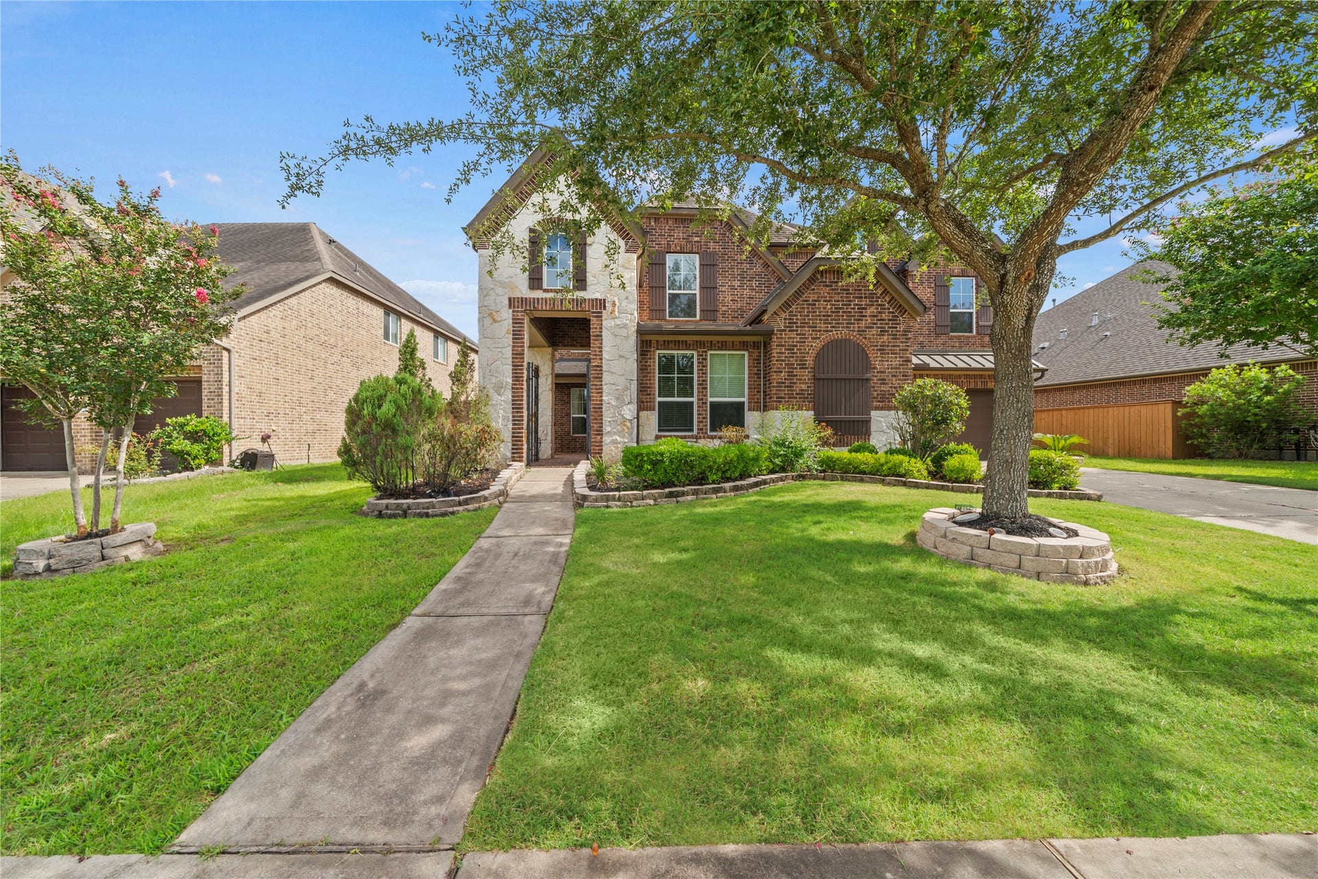 17315 Tomintoul Path, Richmond, TX 77407 A graceful front approach showcases the striking stone-and-brick exterior, lush lawn, and beautifully maintained grounds beneath the canopy of mature trees.