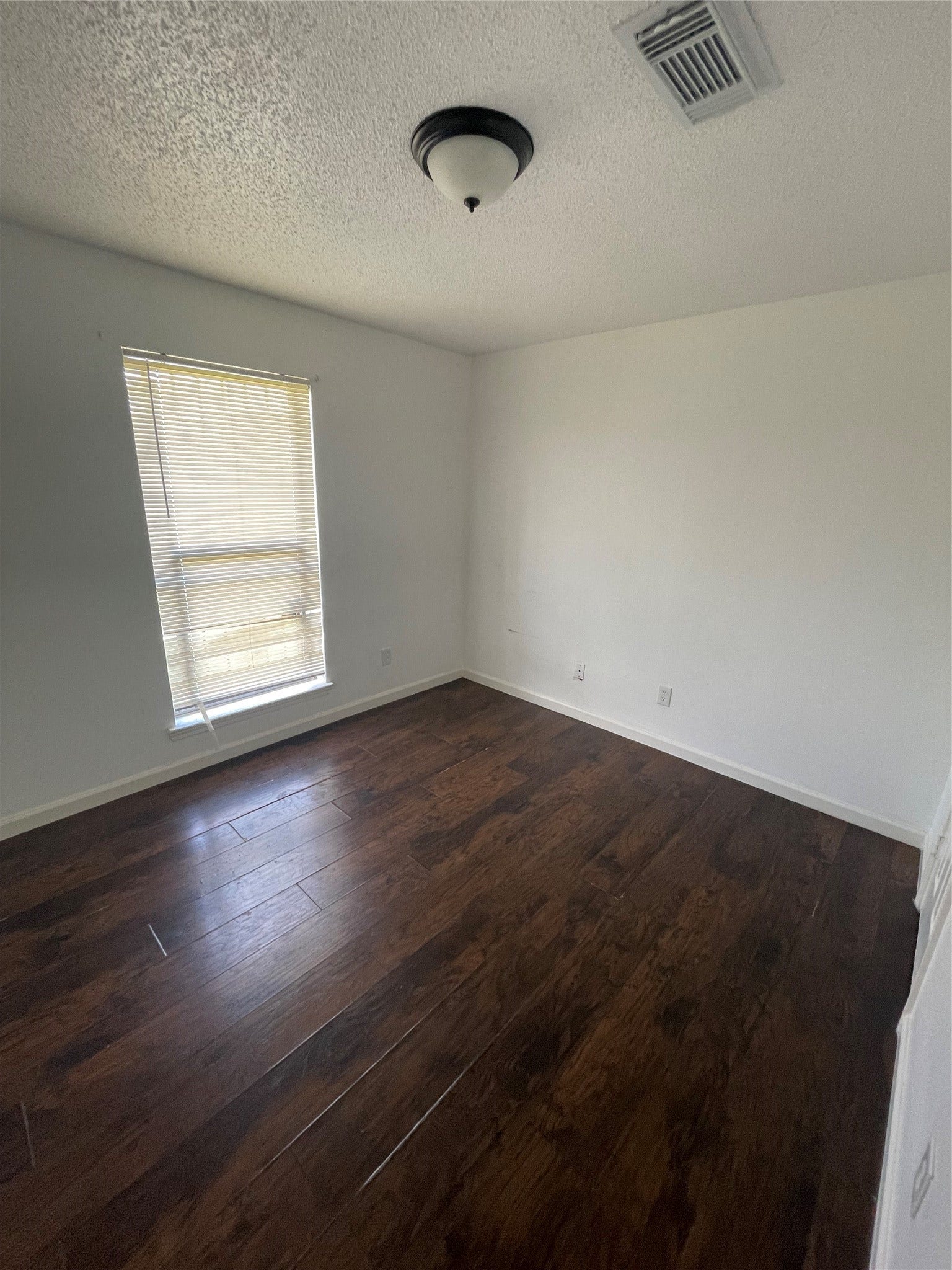 Secondary bedroom featuring rich wood-look flooring, neutral wall colors, a ceiling light fixture, and a window with blinds that allows natural light to fill the space. A versatile room with plenty of possibilities for comfortable living.