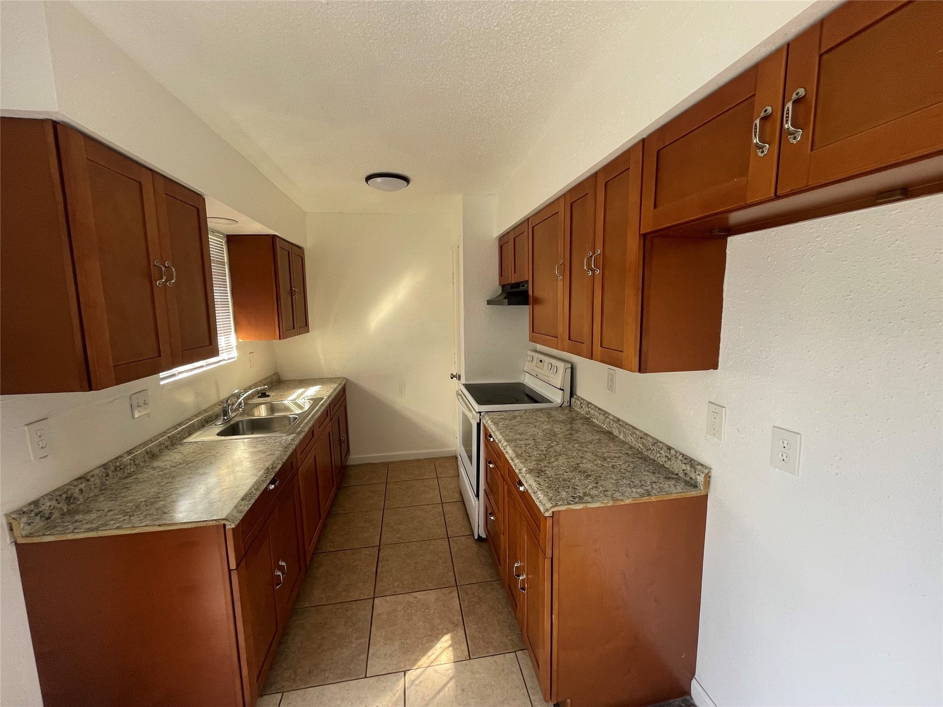 Galley style kitchen featuring warm wood cabinetry, ample cabinet storage, generous countertop space, a double stainless steel sink, electric range, and tile flooring. A window above the sink provides natural light, creating a bright and functional cooking space.