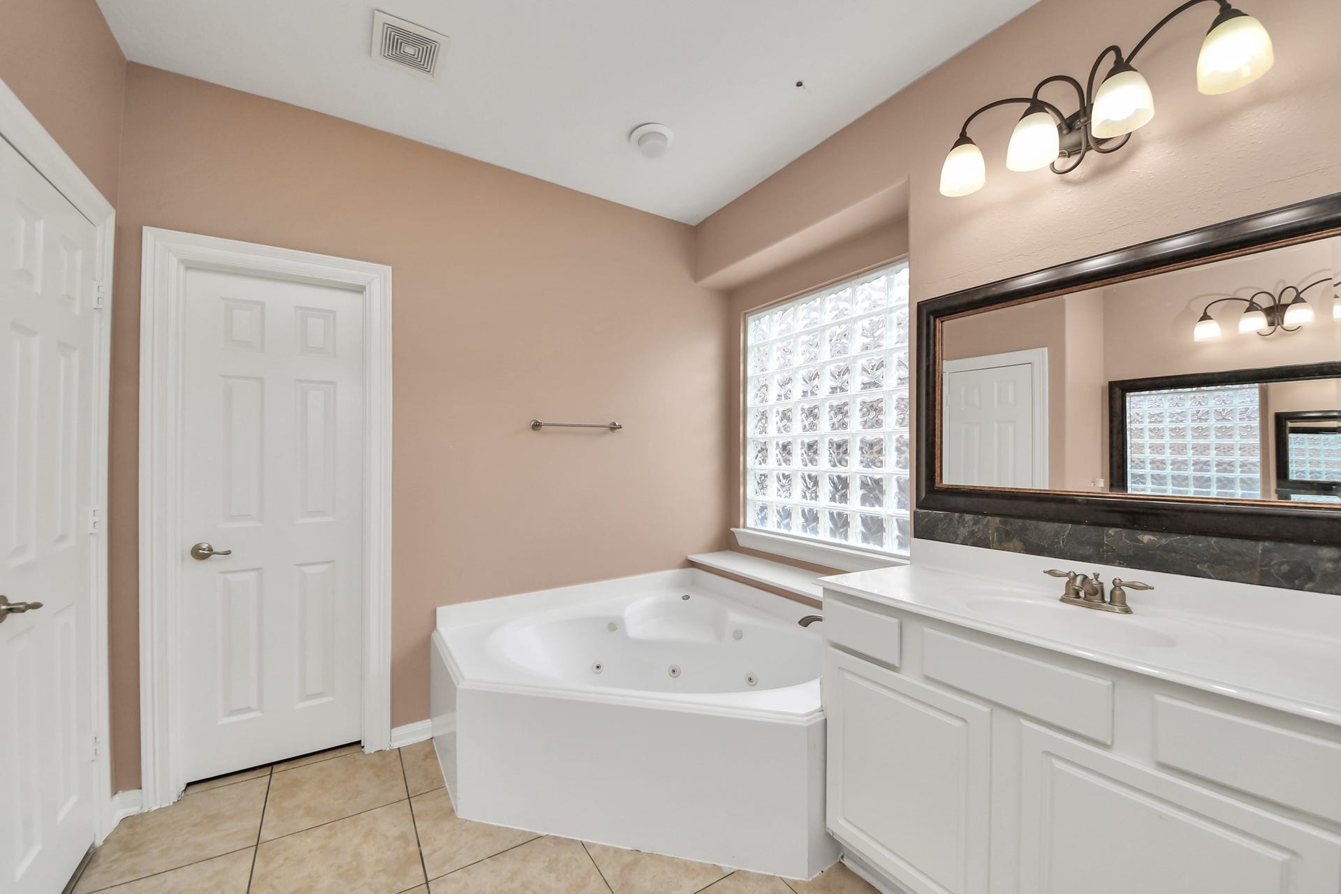 Spacious bathroom featuring a luxurious corner jacuzzi tub, elegant vanity with a large mirror, and soft, natural light from a glass block window.