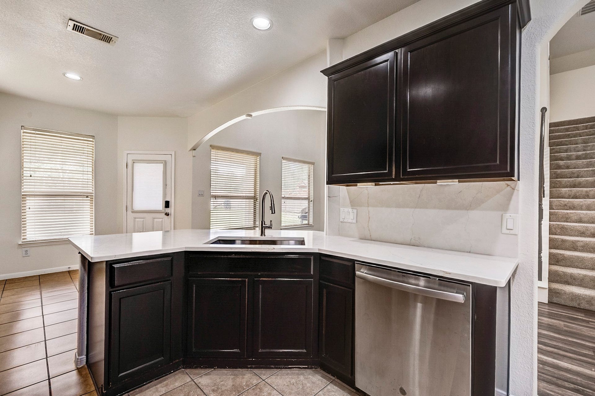 33227 Greenfield Forest Drive, Magnolia, TX 77354 Elegant kitchen featuring dark cabinetry, a sleek stainless steel dishwasher, and a spacious countertop with a modern sink, complemented by abundant natural light from large windows and convenient access to the stairway.