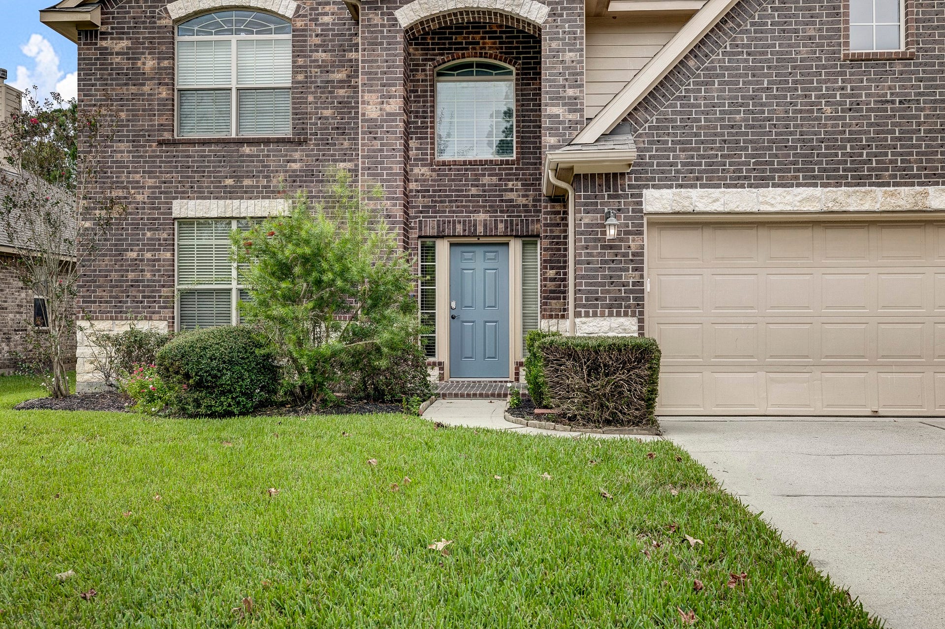 33227 Greenfield Forest Drive, Magnolia, TX 77354 Charming two-story brick home with a welcoming blue front door, complemented by a neatly trimmed lawn and well-maintained shrubs.
