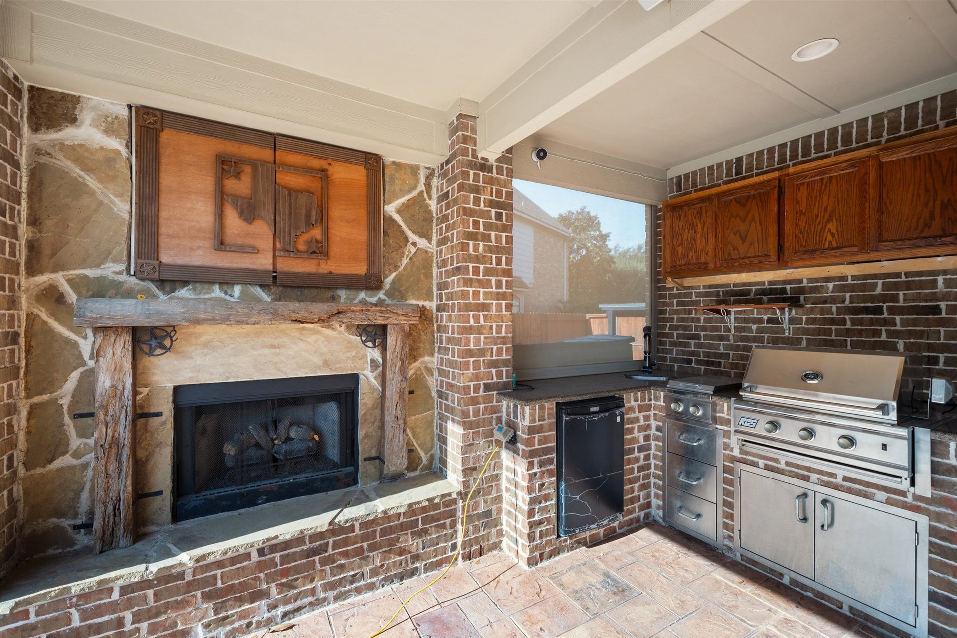 14318 Red Creek Cove Lane, Humble, TX 77396 Outdoor kitchen features a built-in grill, stainless steel appliances, and rustic brickwork. It includes a fireplace with a stone surround, TV in protected cabinet above the fireplace, wooden cabinetry, and a small counter space, ideal for entertaining.