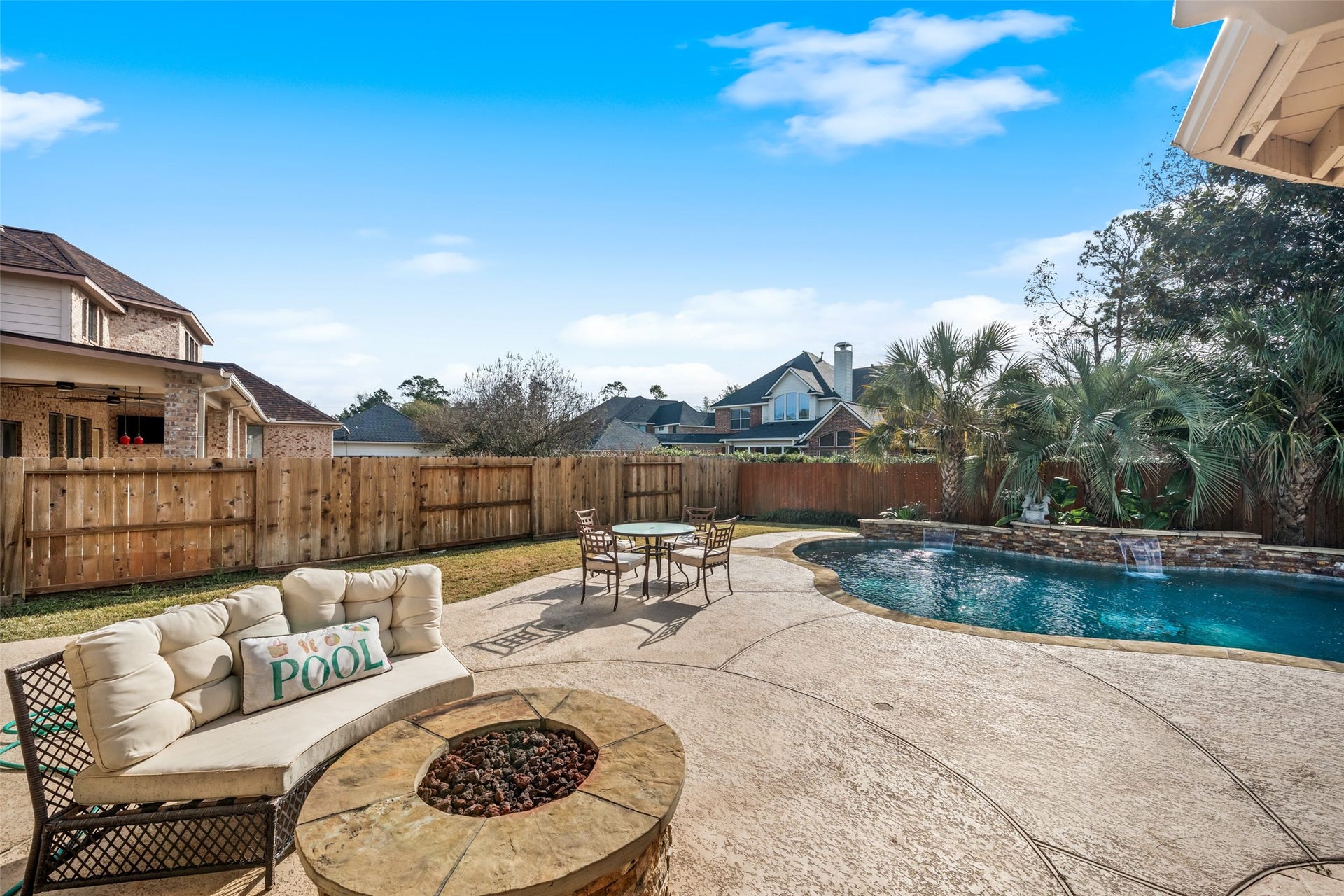 14318 Red Creek Cove Lane, Humble, TX 77396 This backyard features a spacious pool with a waterfall, surrounded by lush greenery and a privacy fence. There's a cozy seating area with a fire pit and a table for outdoor dining, perfect for relaxation and entertaining.