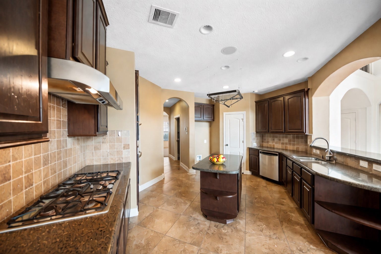 14318 Red Creek Cove Lane, Humble, TX 77396 Kitchen features wood cabinetry, a modern stainless-steel range hood, and a gas cooktop. It has a central island, a double sink, and stainless-steel dishwasher. Arched doorways add a touch of elegance.