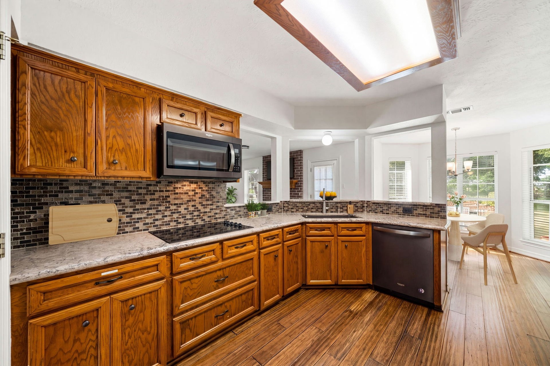 28310 Rose Lane, Katy, TX 77494 Functional kitchen setup showcasing ample wood storage cabinetry, an elegant mosaic tile backsplash, and coordinating appliances including a built-in microwave and dishwasher. The L-shaped workspace flows seamlessly around the durable countertops, complete with an integrated smooth cooktop. Rich wood-look flooring stretches across the room, which opens directly to the dining area via a raised breakfast bar ledge.
