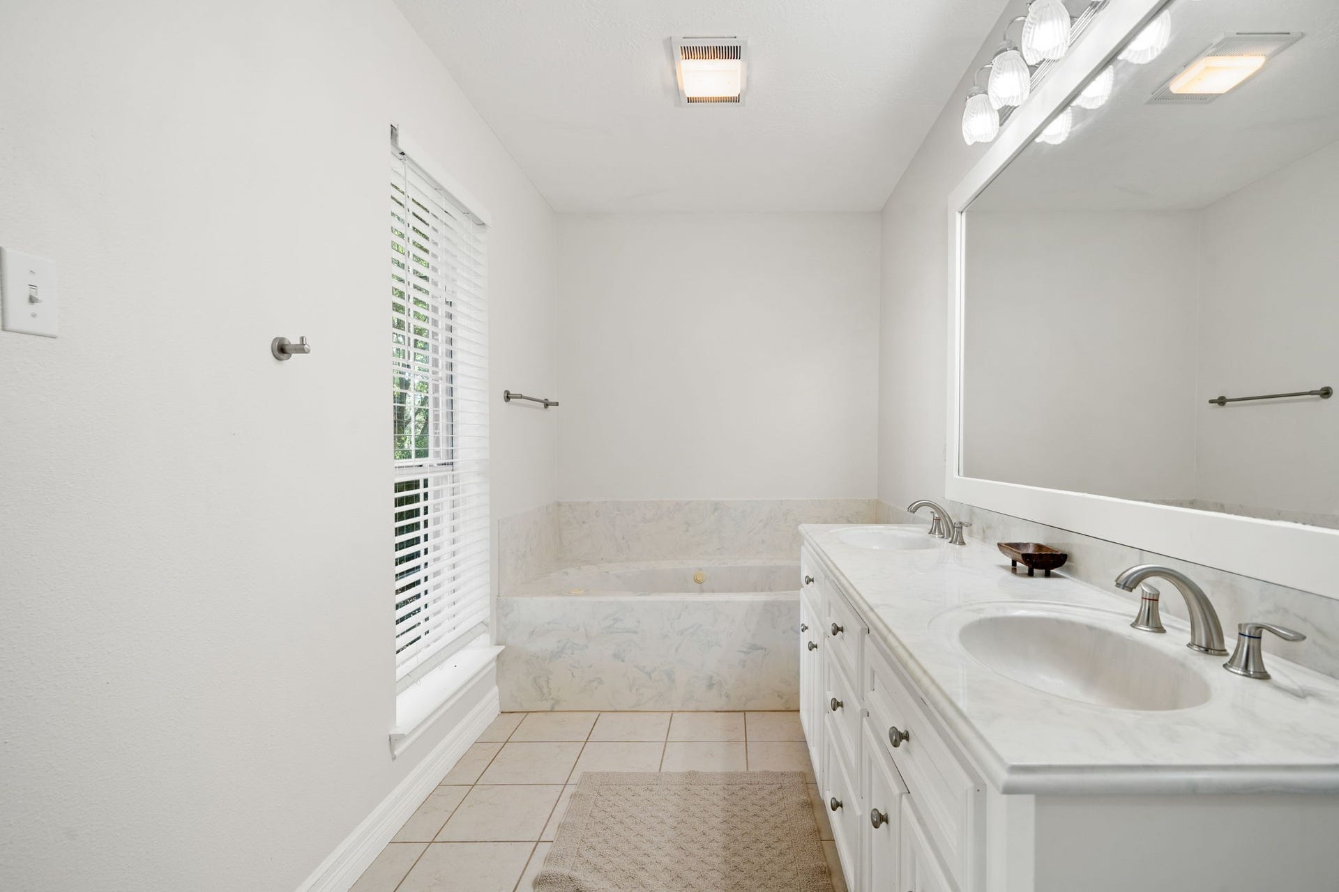28310 Rose Lane, Katy, TX 77494 Bright and airy primary bathroom showcasing a dual-sink vanity with crisp white cabinetry and a spacious, well-lit mirror. A deep tub is positioned perfectly at the far end of the room beneath classic overhead lighting. Large-format tile flooring, a neutral color palette, and a well-placed window fill the entire space with a clean and refreshing ambiance.