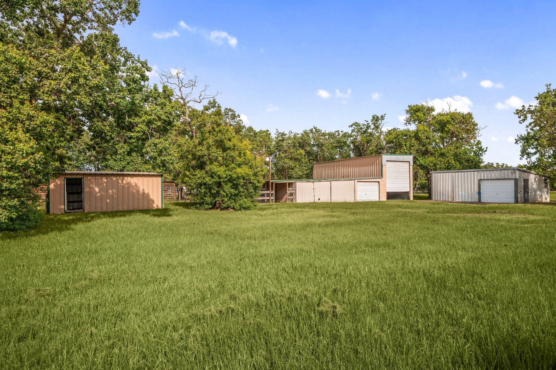 28310 Rose Lane, Katy, TX 77494 Versatile acreage clearing highlighting a cluster of practical outbuildings and storage sheds situated along the edge of a deep tree line. A wide, open expanse of green grass dominates the foreground, providing excellent workspace maneuverability or room to park large equipment and vehicles. Tucked away beneath a vibrant blue sky, this functional section of the property is ideal for agricultural projects, workshops, or heavy-duty storage needs.