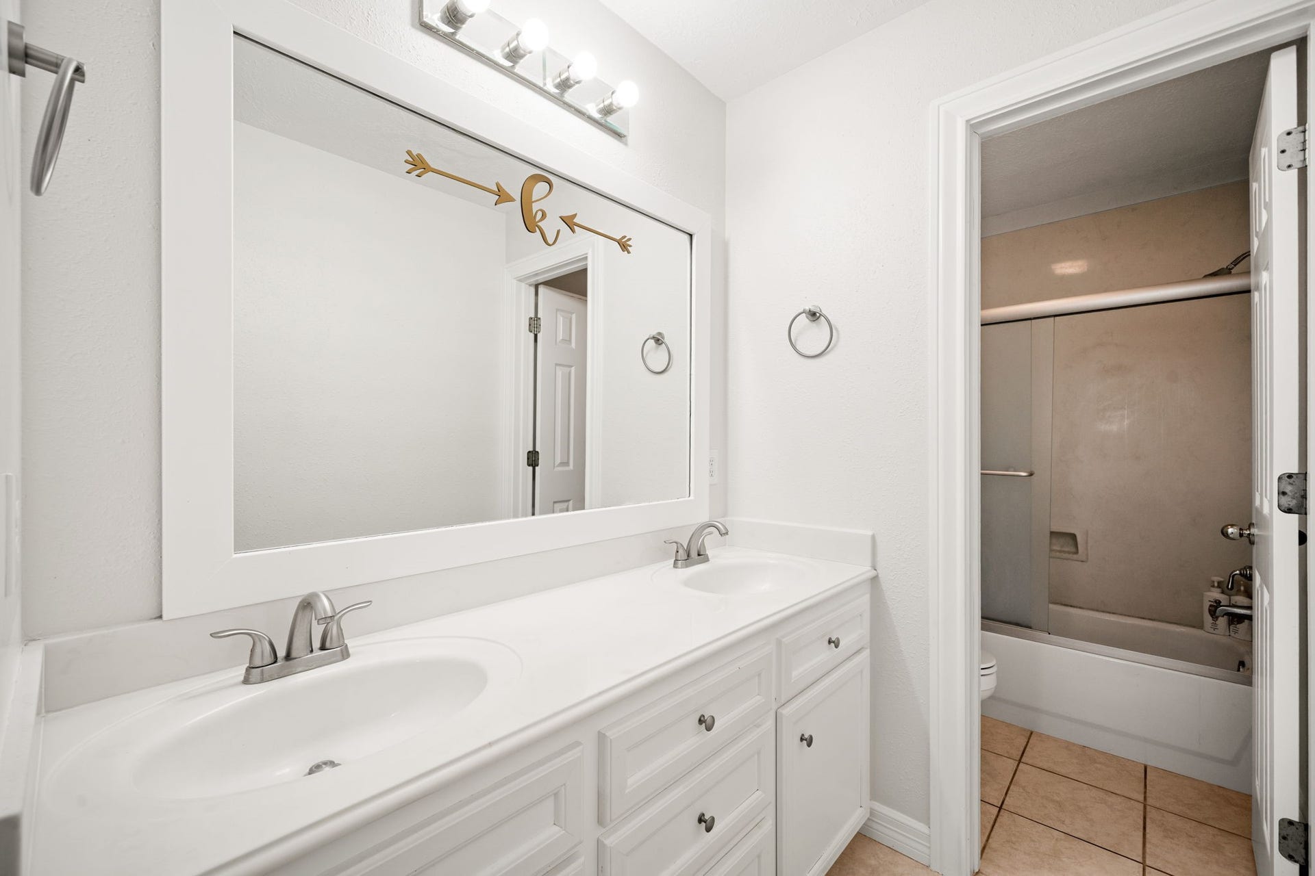 28310 Rose Lane, Katy, TX 77494 Bright and functional full bathroom showcasing a spacious double vanity with crisp white cabinetry and sleek undermount sinks. A large white-framed mirror anchors the wall, beautifully illuminated by modern multi-light vanity fixtures. Large-format tile flooring transitions effortlessly through an open doorway into a separate room featuring a classic tub and shower combination.