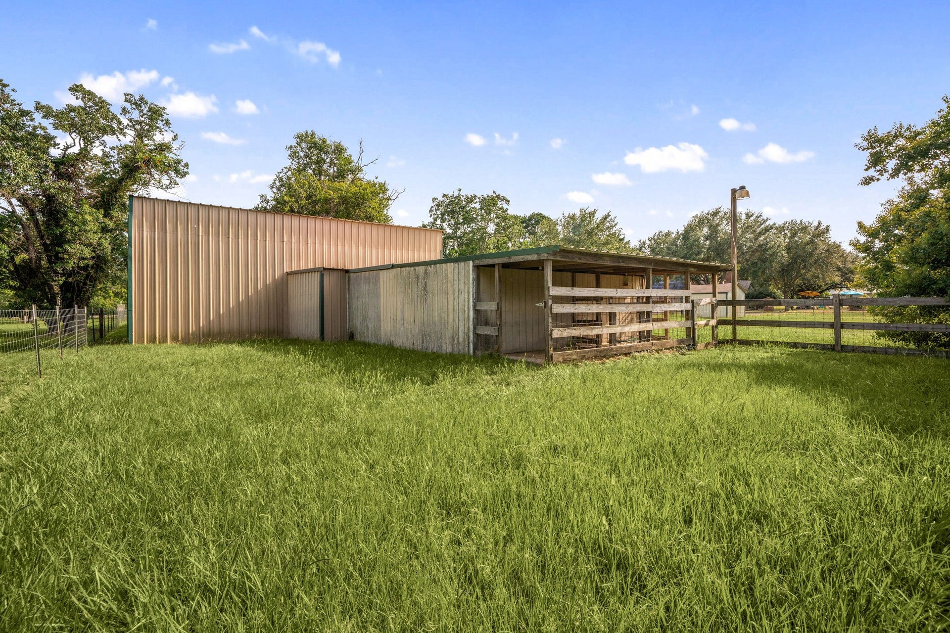 28310 Rose Lane, Katy, TX 77494 Dedicated livestock structure featuring an open-sided run-in shelter integrated with robust timber fencing panels. The paddock layout sits on a gentle grassy clearing, surrounded by a backdrop of mature trees and clear blue skies, offering excellent shelter for animals or versatile farm storage. A secure gate provides easy entry and pasture management, making this a prime feature for equestrian buyers or hobby farmers.