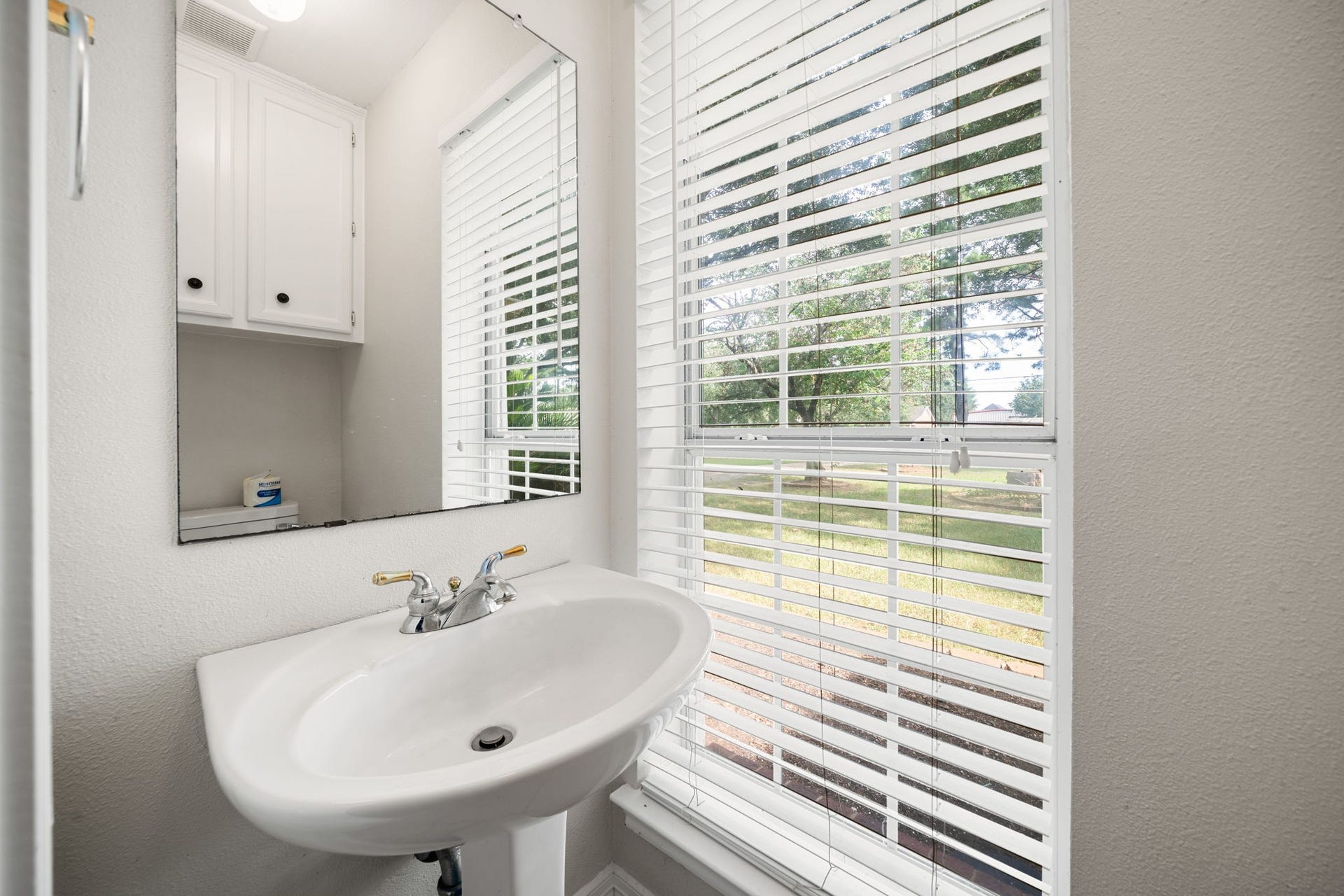 28310 Rose Lane, Katy, TX 77494 Convenient half bathroom featuring a classic white pedestal sink and a large wall mirror. A bright window equipped with adjustable horizontal blinds fills the space with natural light while providing view control. Crisp white cabinetry is mounted above the toilet location, maximizing vertical storage in this highly functional layout positioned off the attached garage.