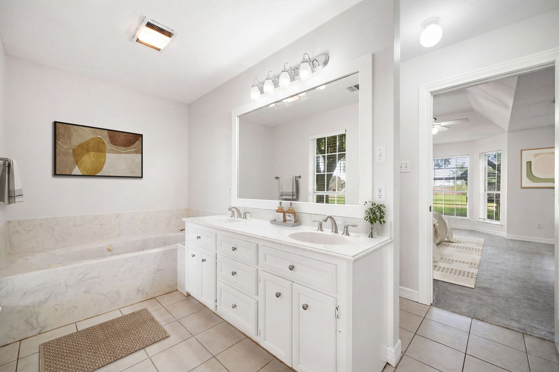 28310 Rose Lane, Katy, TX 77494 Spa-like primary bathroom featuring a long double vanity with crisp white cabinetry and a large framed mirror illuminated by modern vanity lighting. A deep soaking tub sits adjacent to the vanity, providing a perfect spot for relaxation. Large-format tile flooring coordinates seamlessly with the bright color palette, while an open doorway offers a convenient transition directly into the primary bedroom.