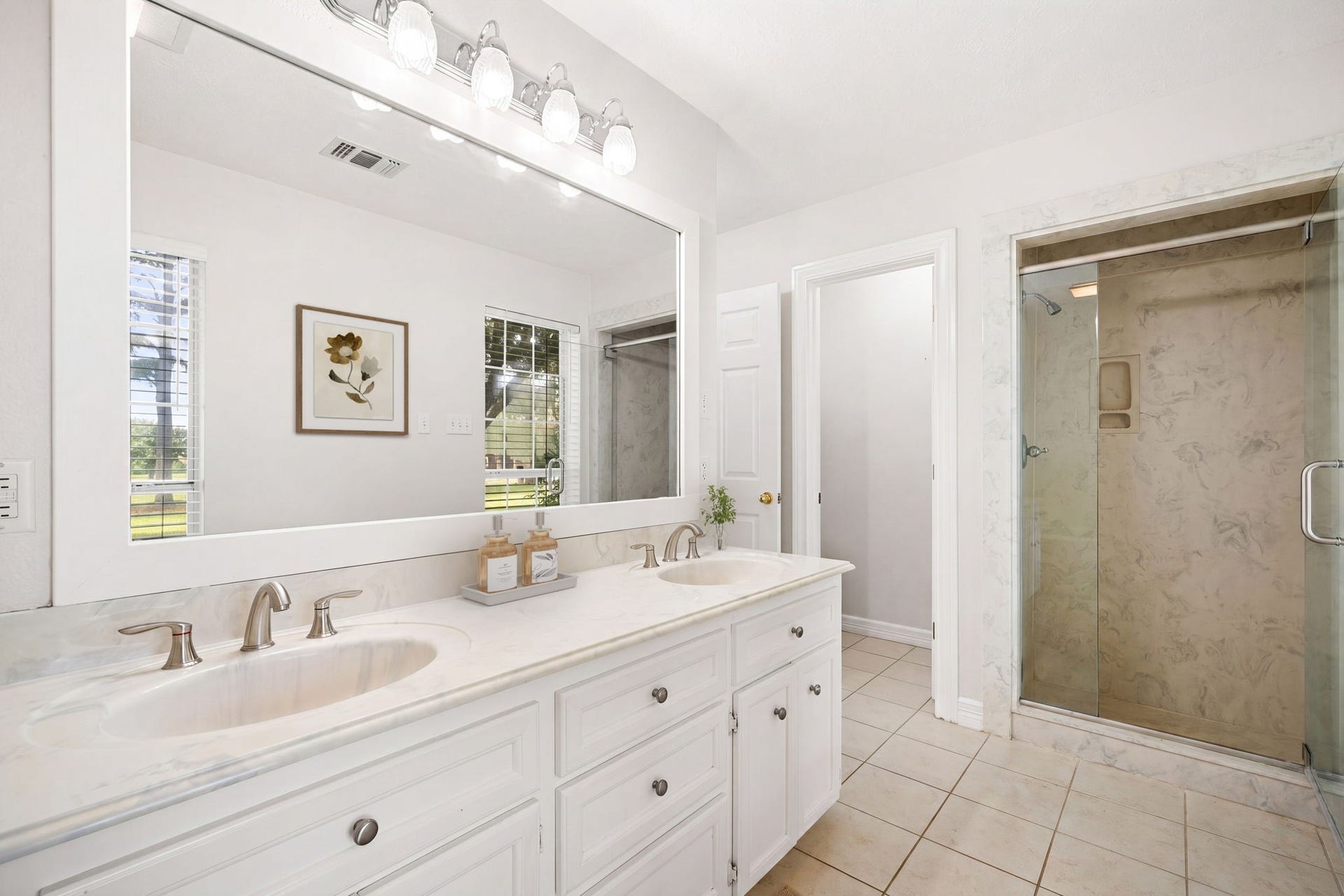 28310 Rose Lane, Katy, TX 77494 Bright, modern en-suite bathroom showcasing an expansive double vanity with a sleek white countertop and undermount sinks. A large, white-framed mirror spans the vanity area, illuminated by a contemporary multi-light vanity fixture. The space is completed by crisp white cabinetry offering ample storage, large-format tile flooring, and a separate walk-in shower enclosed by a glass door.