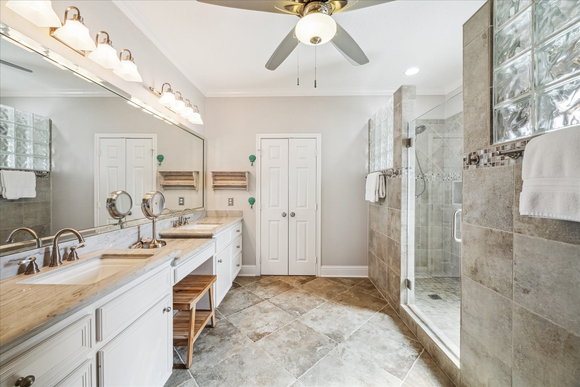 519 Westerly Drive, Tiki Island, TX 77554 The primary bathroom features a spacious layout with a double vanity, large mirrors, and ample lighting. It includes a double walk-in shower with glass block accents and neutral tile flooring. A ceiling fan adds comfort, and theres a sleek, modern design throughout.