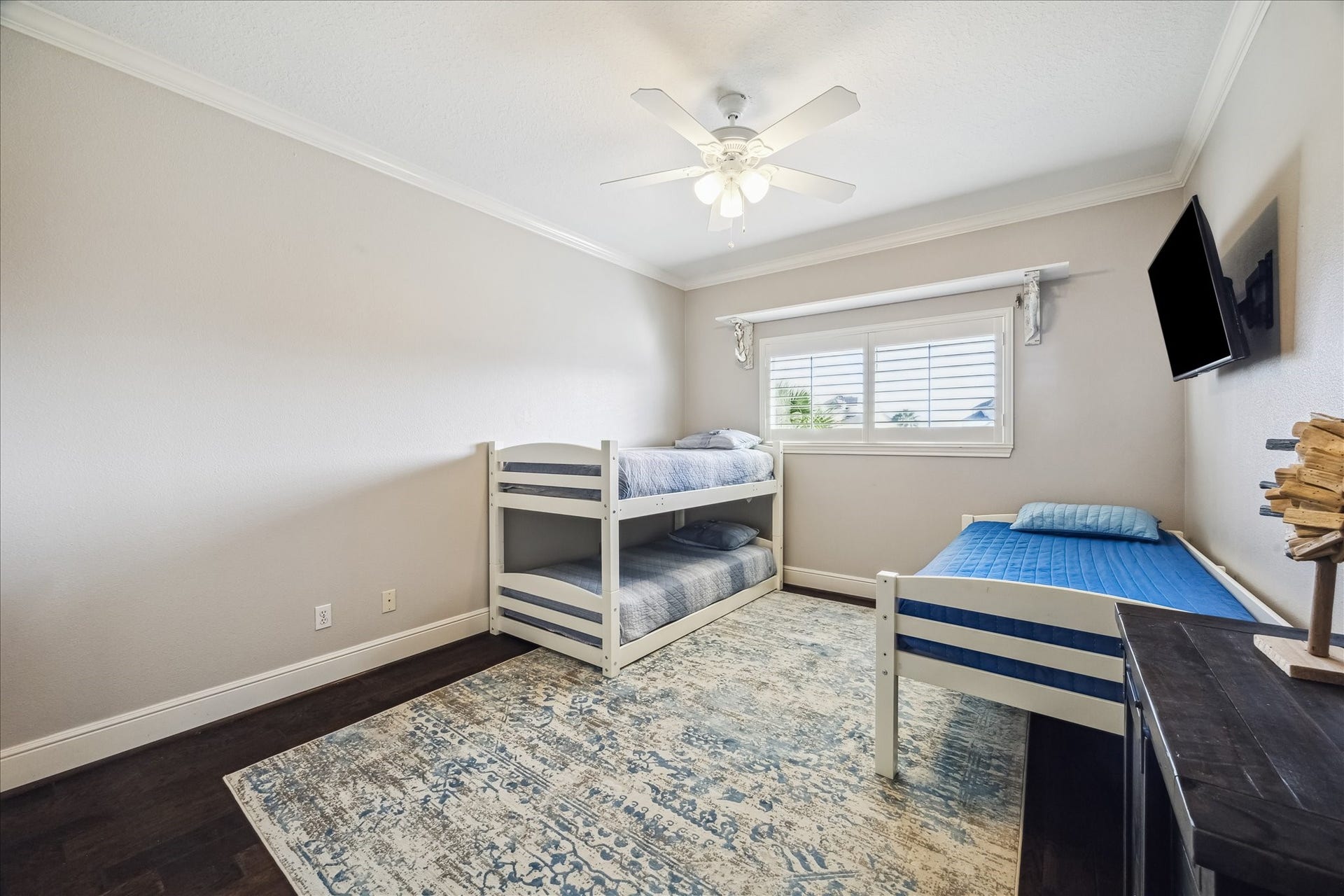 519 Westerly Drive, Tiki Island, TX 77554 This secondary bedroom features a clean, modern design with neutral walls, dark hardwood floors, and a ceiling fan. It includes a bunk bed and a single bed, with a large window for natural light. A wall-mounted TV offers entertainment, and a patterned area rug adds warmth and style.