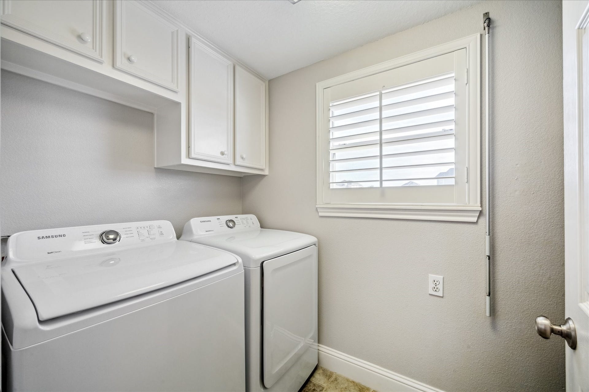 519 Westerly Drive, Tiki Island, TX 77554 This laundry room is bright and functional, featuring a washer and dryer set, white cabinetry for storage, and a window with shutters for natural light.