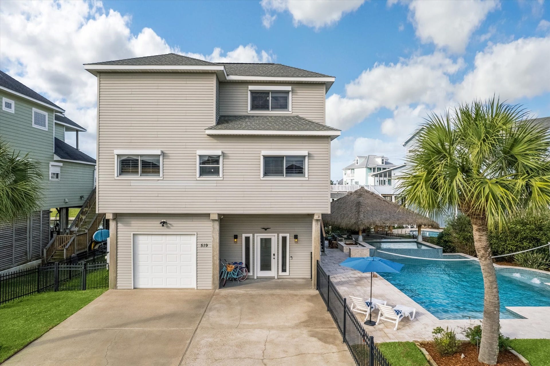 519 Westerly Drive, Tiki Island, TX 77554 This modern, elevated home features a spacious driveway, a garage, and a luxurious entertaining space with a large pool and patio area. The property is accented by palm trees, offering a tropical feel.