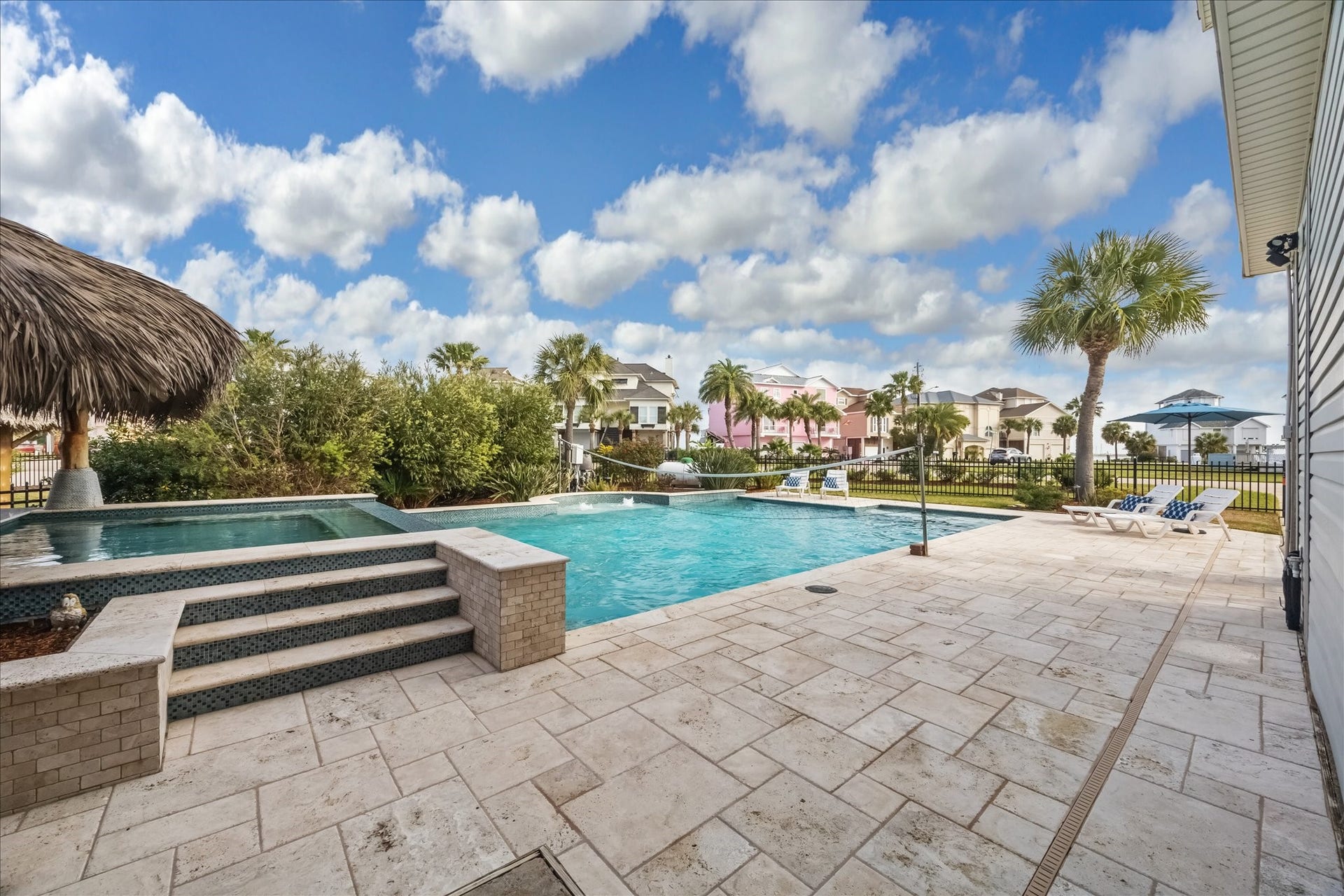 519 Westerly Drive, Tiki Island, TX 77554 Luxurious pool, a stone-tiled deck, and a palapa. It features lush greenery, palm trees, and a view of colorful neighboring houses, creating a tropical oasis feel.