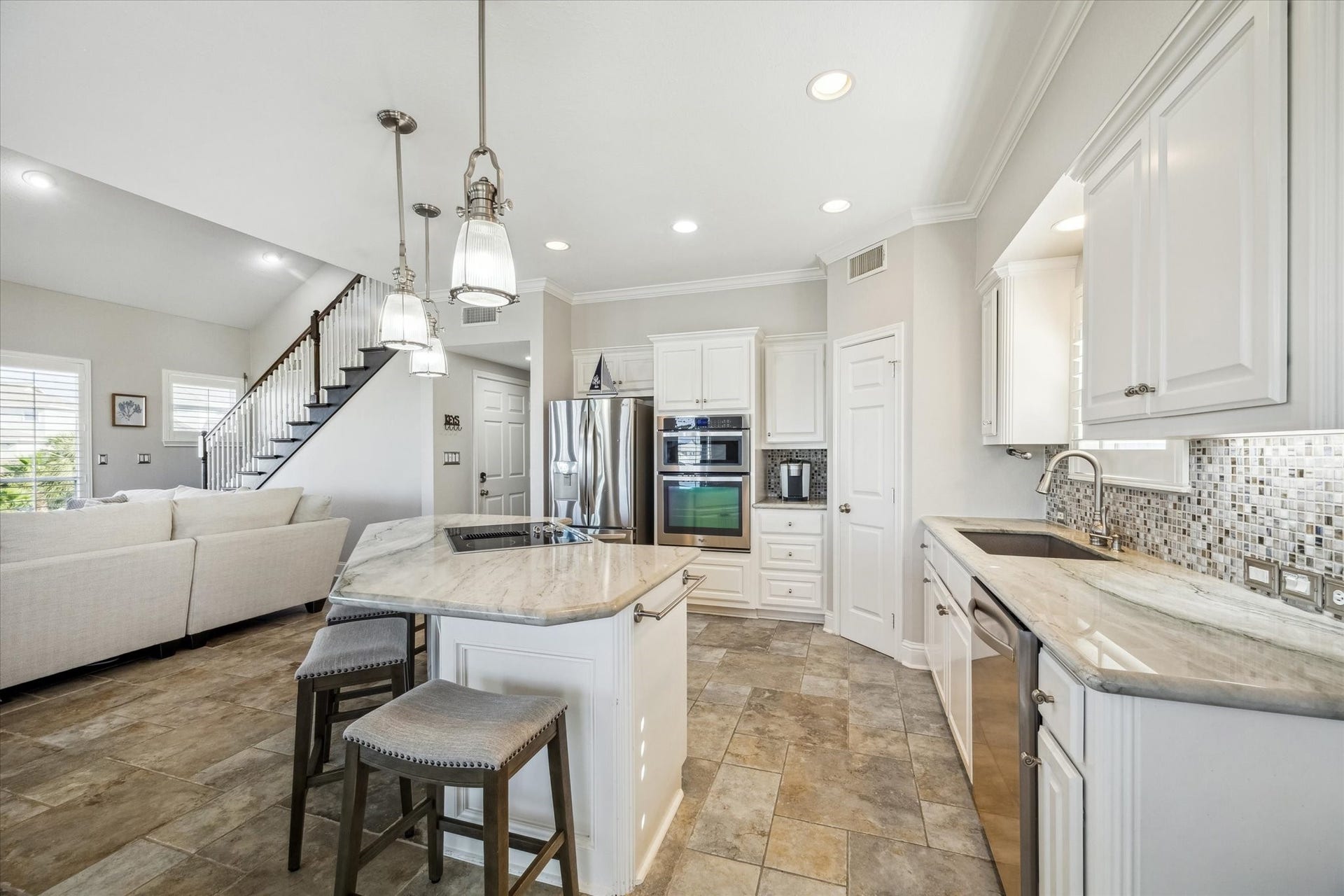 519 Westerly Drive, Tiki Island, TX 77554 Bright, open-concept kitchen and living area. The kitchen features modern stainless steel appliances, white cabinetry, and a stylish tile backsplash. A central island offers additional seating and workspace. Adjacent is a cozy living area with a staircase leading upstairs, creating a welcoming and functional space.