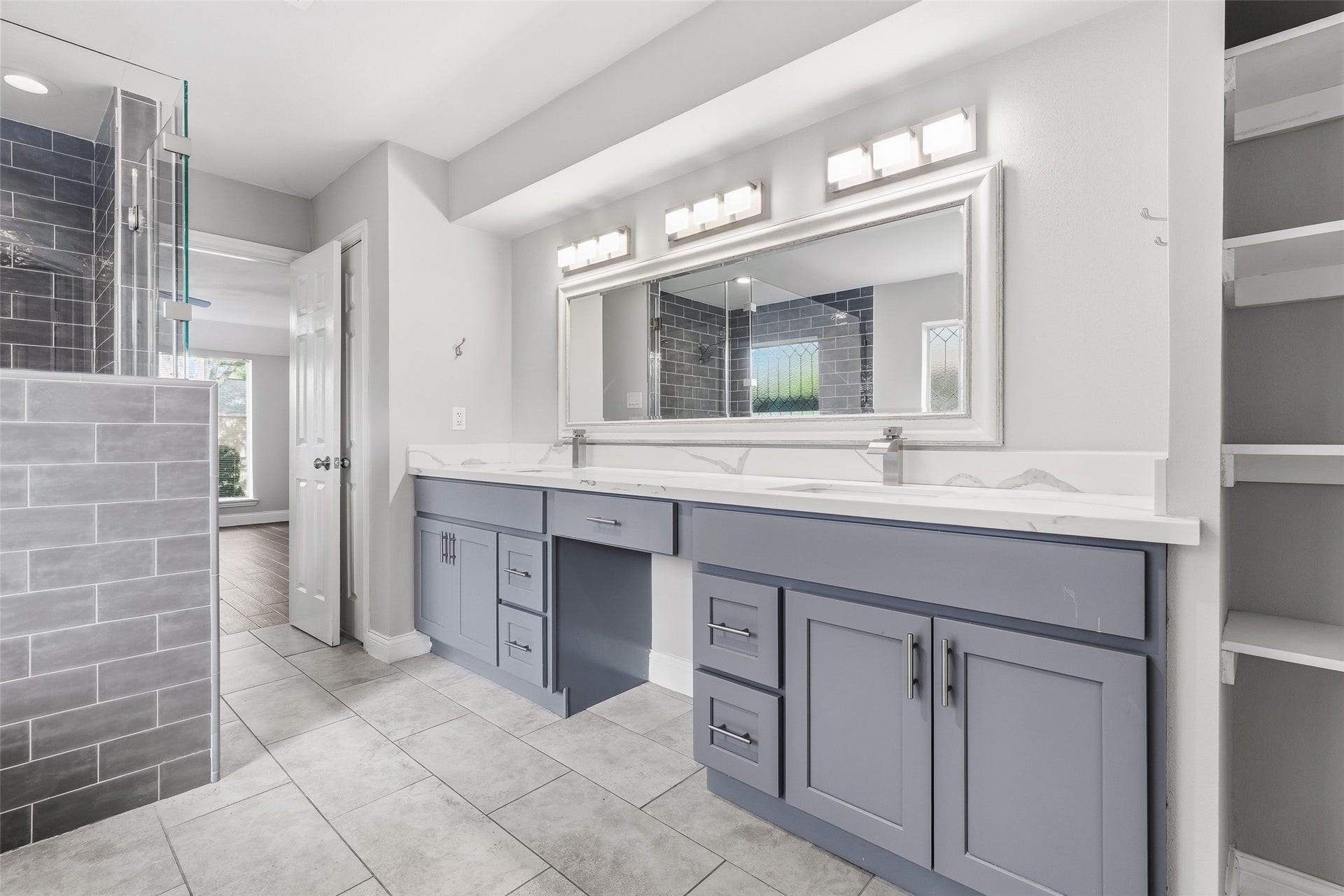 Spacious bathroom with a modern double vanity, sleek gray cabinetry, and a large mirror. The room features elegant tiled flooring and a glass-enclosed shower with contemporary gray tiles.