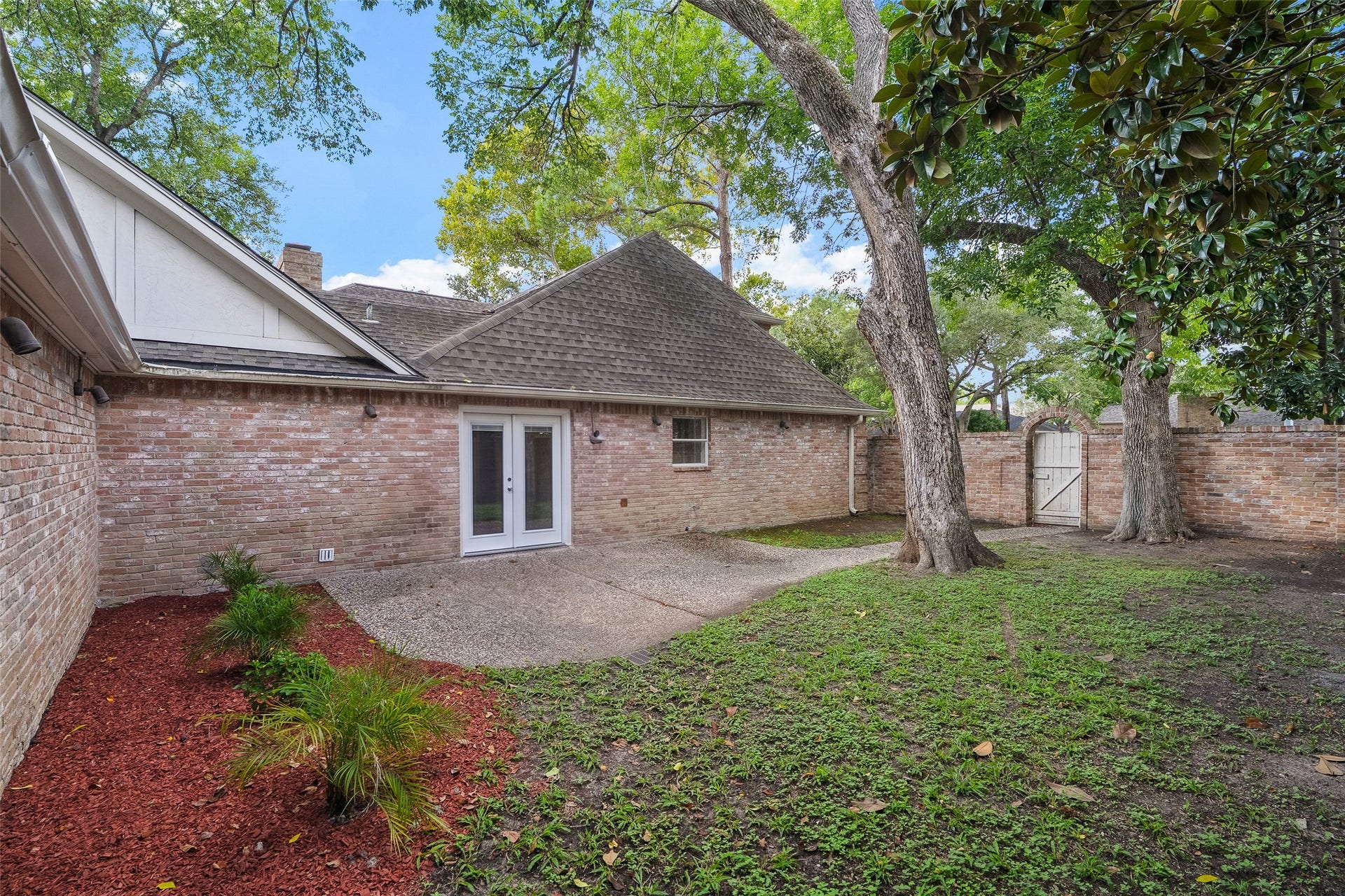 Charming brick exterior with French doors opening to a spacious backyard, featuring mature trees and a private gate, perfect for outdoor relaxation.