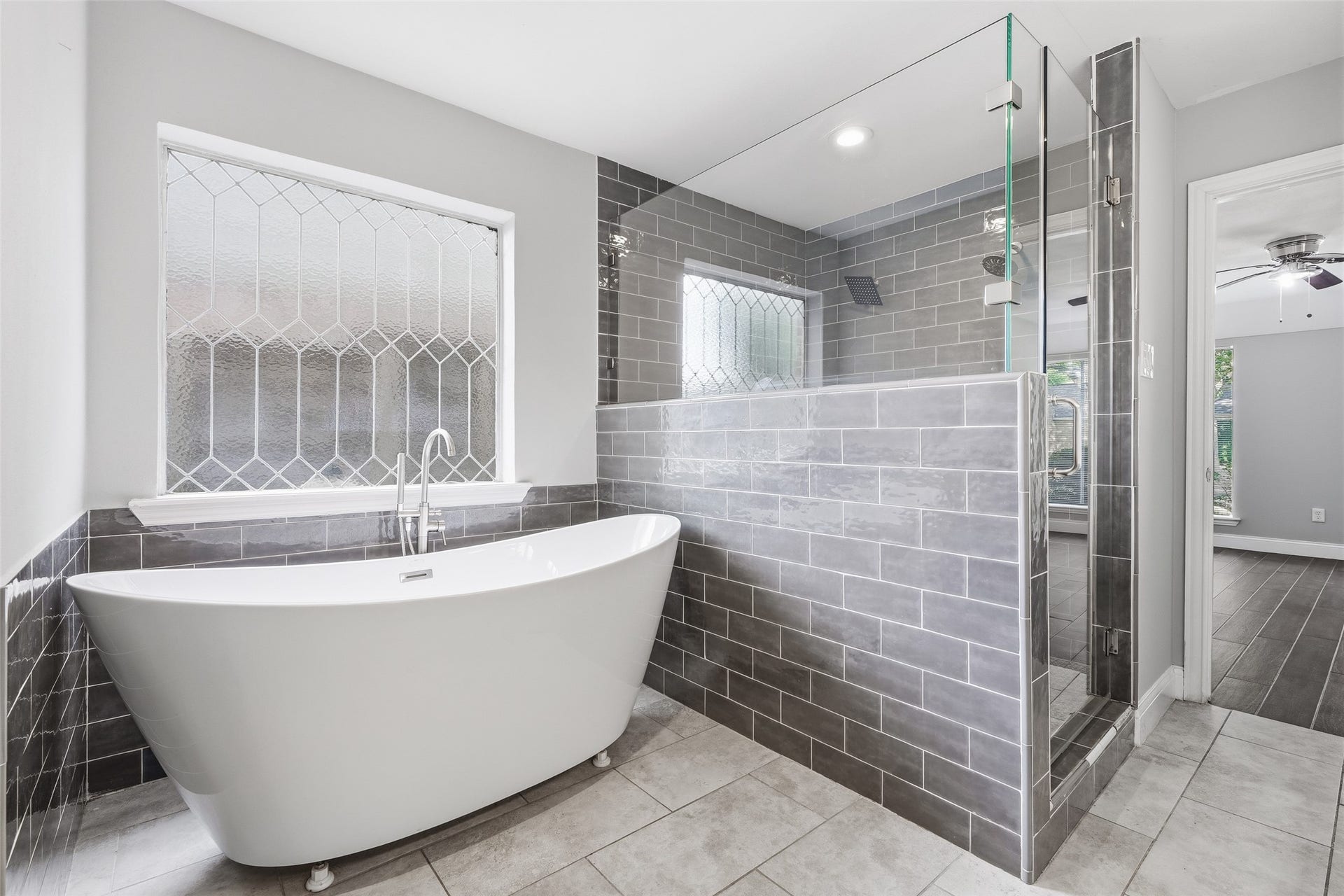 Experience luxury in this elegant bathroom featuring a freestanding tub, modern glass-enclosed shower, and stylish gray tile accents, complemented by a beautiful window.
