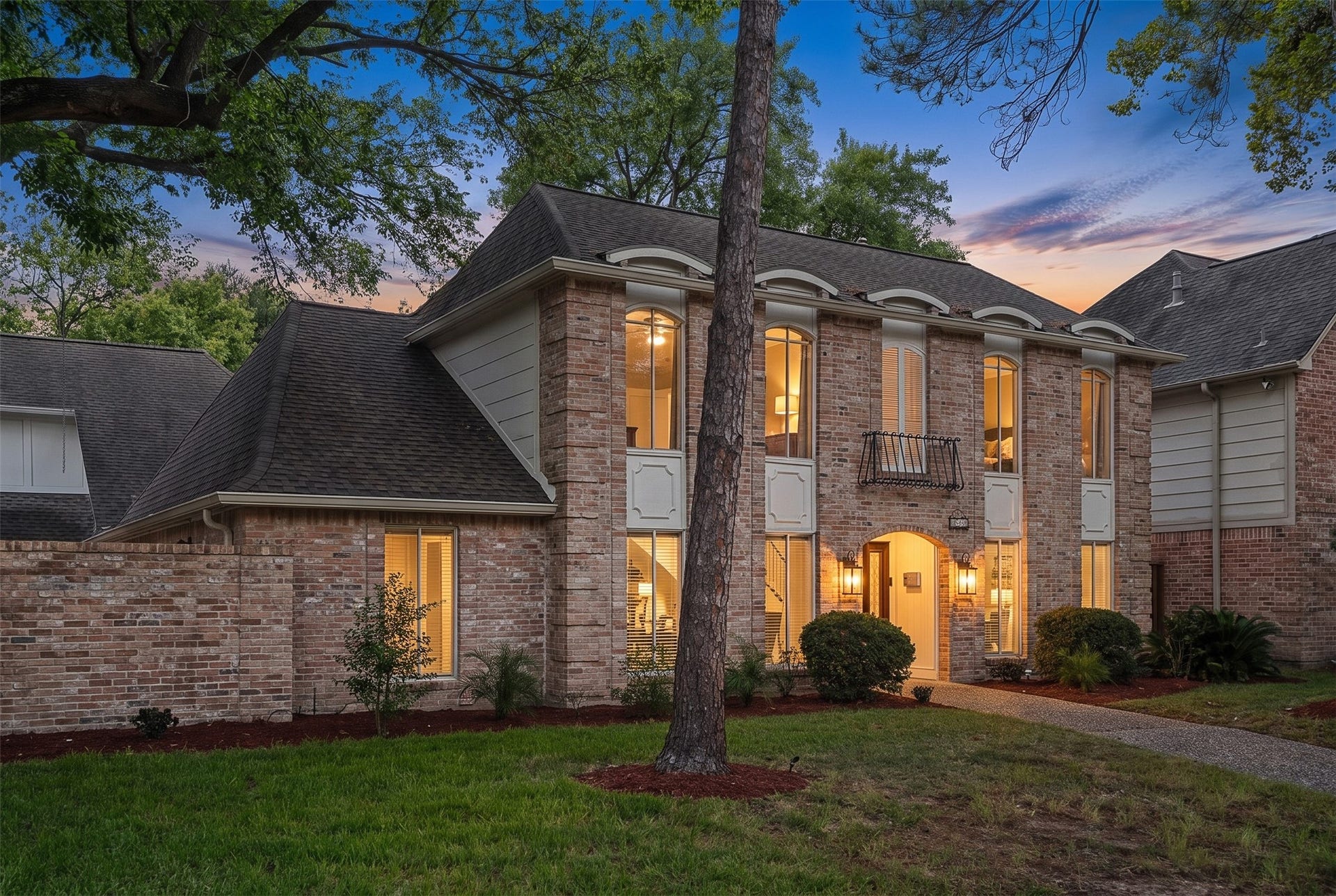 Elegant two-story brick home with large windows and charming architectural details, nestled in a lush, tree-lined neighborhood.