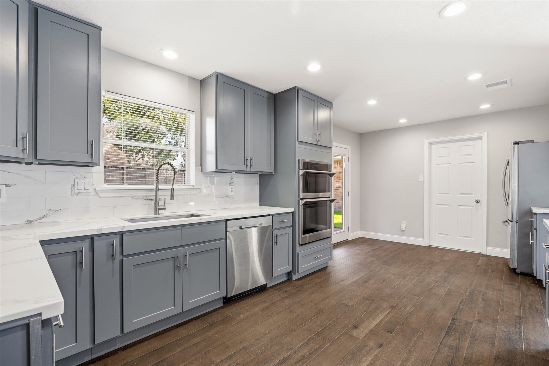 Sleek modern kitchen featuring elegant gray cabinetry, stainless steel appliances, and pristine white countertops, all complemented by rich dark flooring and abundant natural light.