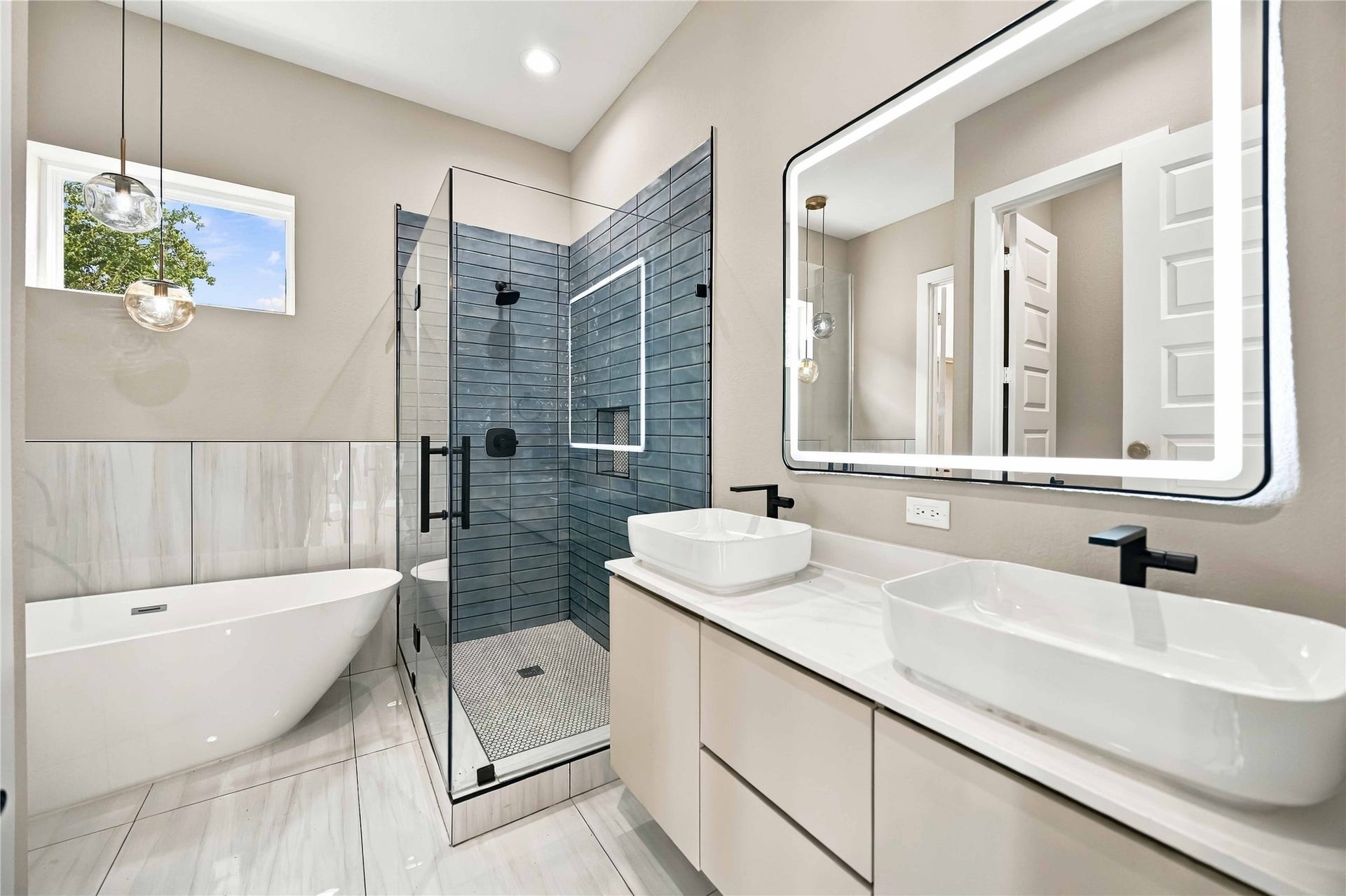 3503 Rebecca Street, #E/F, Houston, TX 77021 Spa-inspired primary bathroom featuring a freestanding soaking tub, glass-enclosed walk-in shower with contemporary blue-gray tile, dual vessel sinks, a sleek white vanity with light countertops, and a striking illuminated mirror. Matte black fixtures and elegant pendant lighting add a sophisticated modern finish.