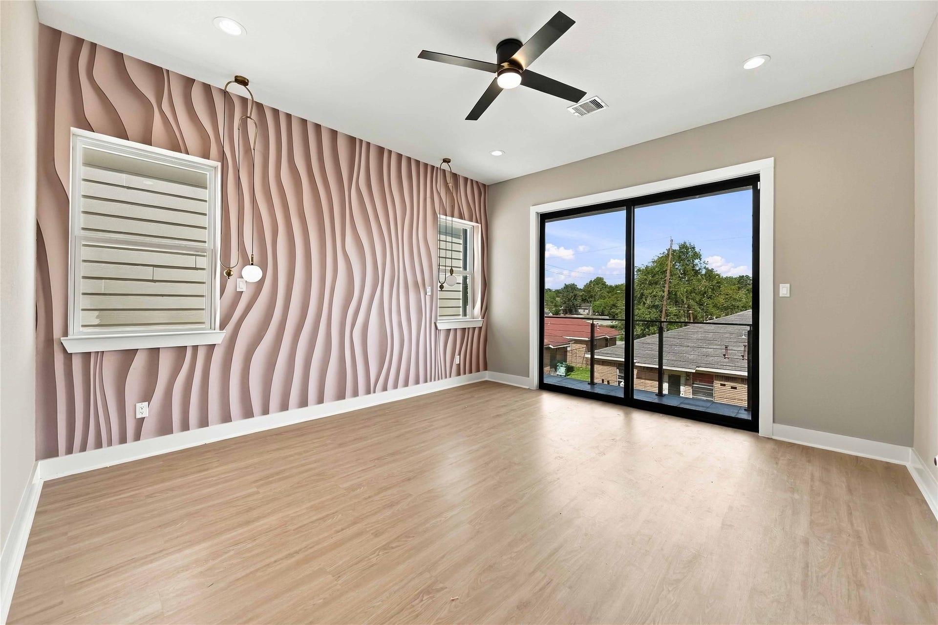 3503 Rebecca Street, #E/F, Houston, TX 77021 Contemporary primary bedroom highlighted by a distinctive sculptural blush-toned accent wall, warm wood-look flooring, modern pendant lighting, and a sleek ceiling fan. Expansive, black-framed sliding glass doors provide abundant natural light and access to a private balcony, creating a stylish and inviting retreat.